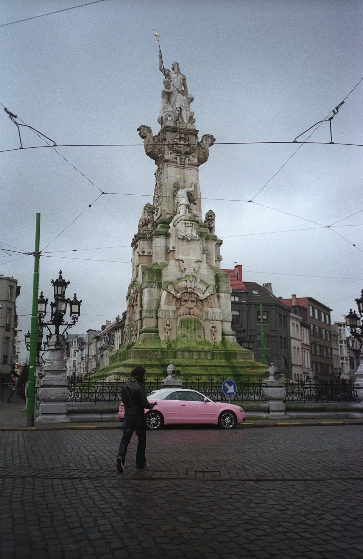 A historic stone monument with elaborate carvings and statues, topped with a statue of a man holding a staff, located in an urban square with cobblestone pavement, lampposts, and surrounding buildings, under an overcast sky.