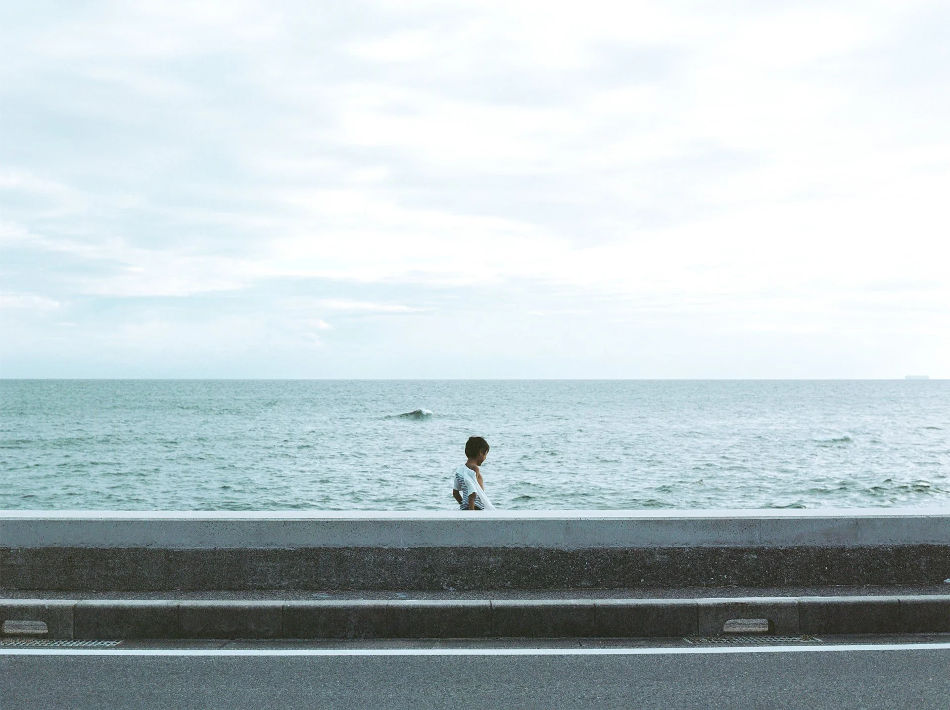 A boy walking along the shore of the ocean with mountains in the background, seen from a road with a guardrail.