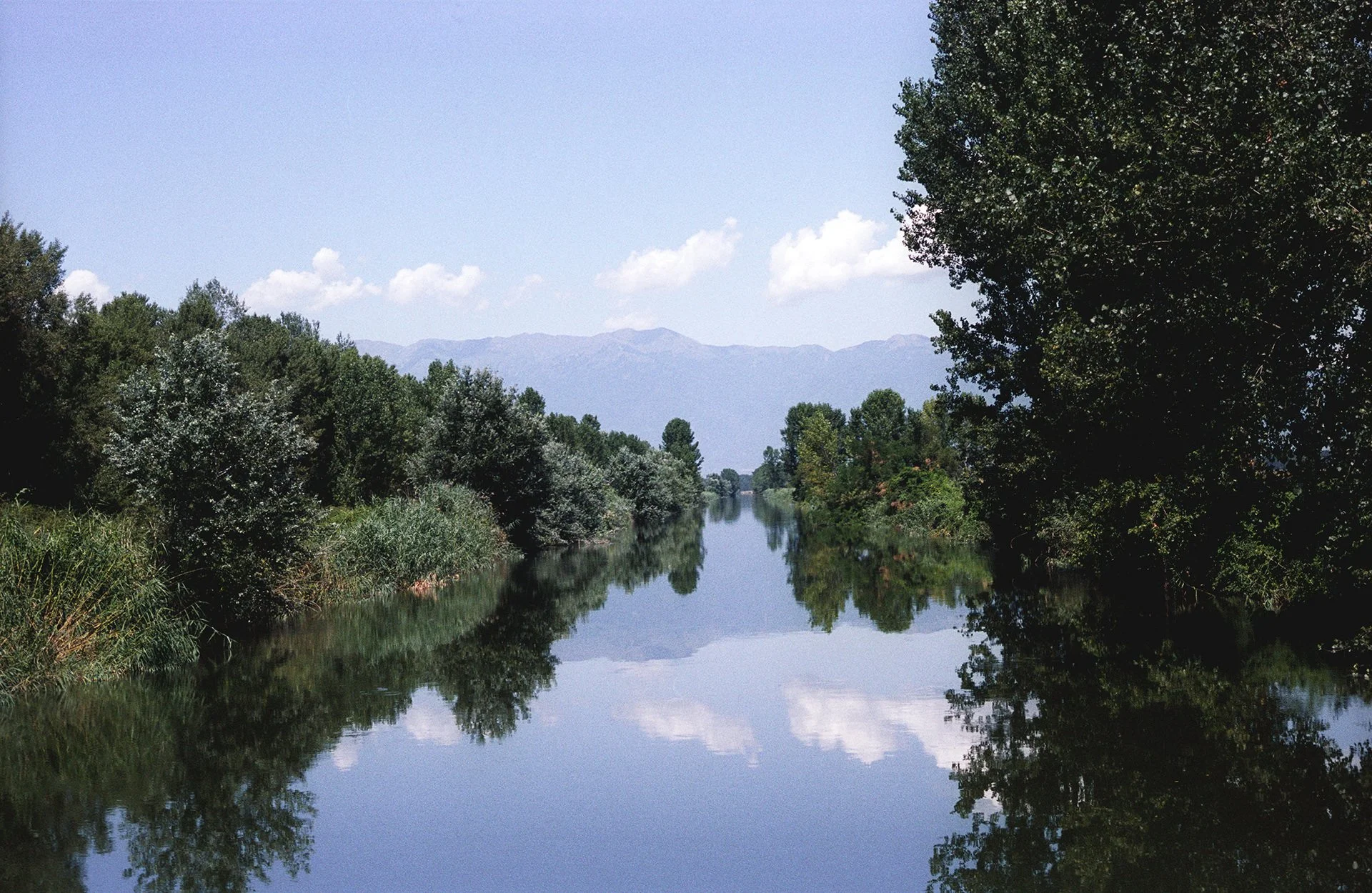 A calm river flanked by lush green trees on both sides, with mountains in the distance and a clear blue sky with a few clouds.