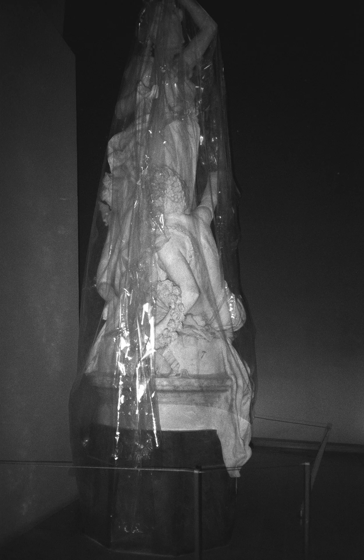 A sculpture of a woman dressed in historical attire, protected by a clear plastic covering, on display in a gallery.
