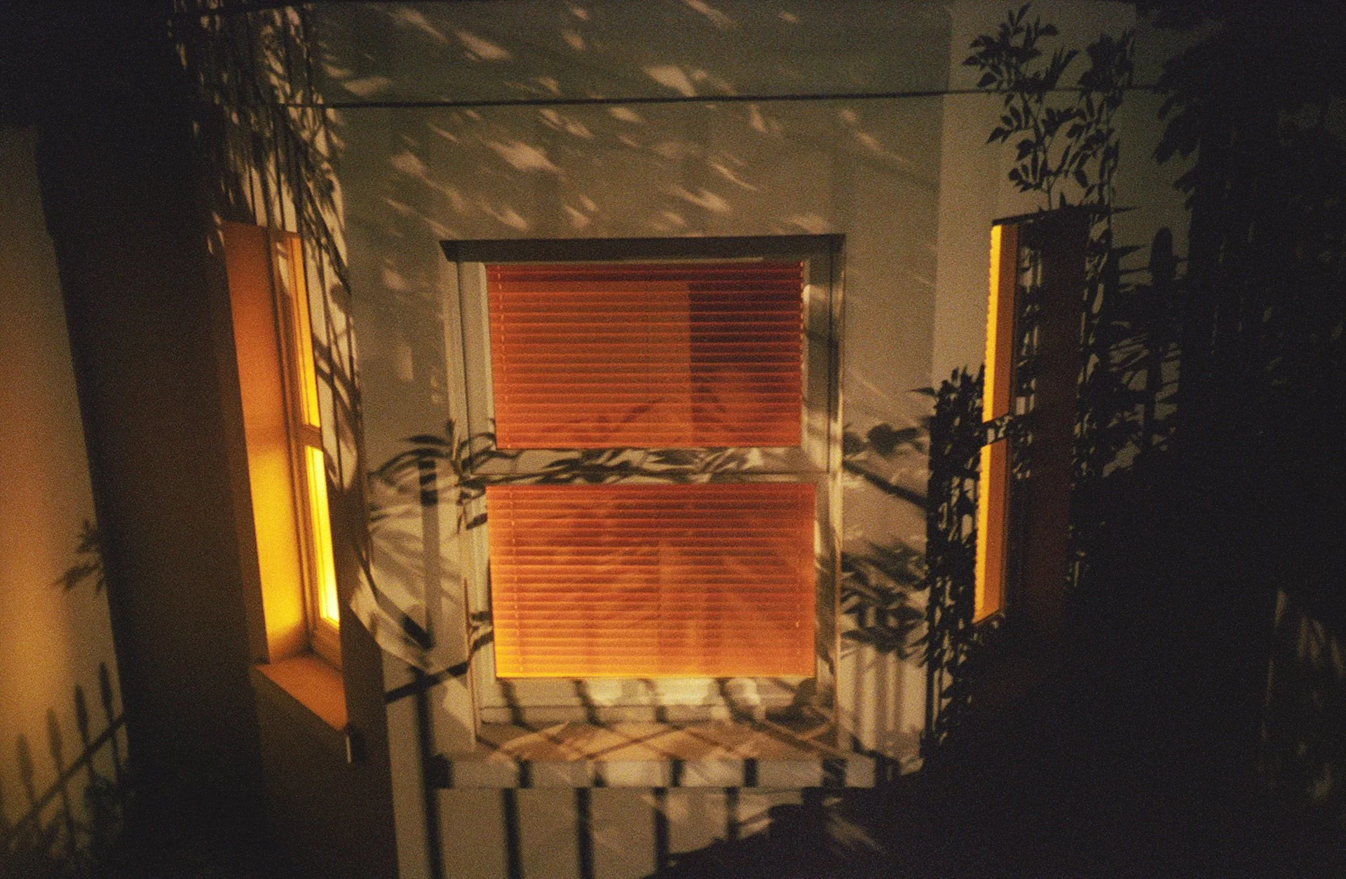 Close-up of a house window with closed wooden blinds, surrounded by decorative black metal grid shadows, illuminated by warm yellow lighting.