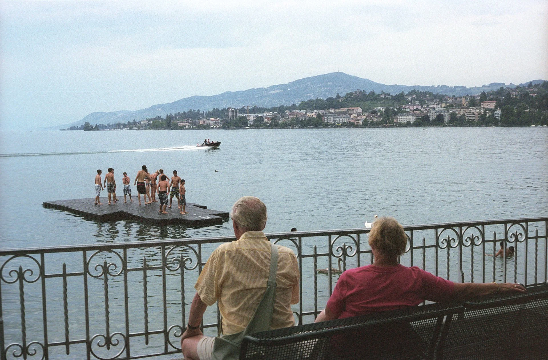 People sitting on a bench by a lake, watching children in swim trunks jumping from a dock into the water, with a boat passing by and a cityscape with mountains in the background.