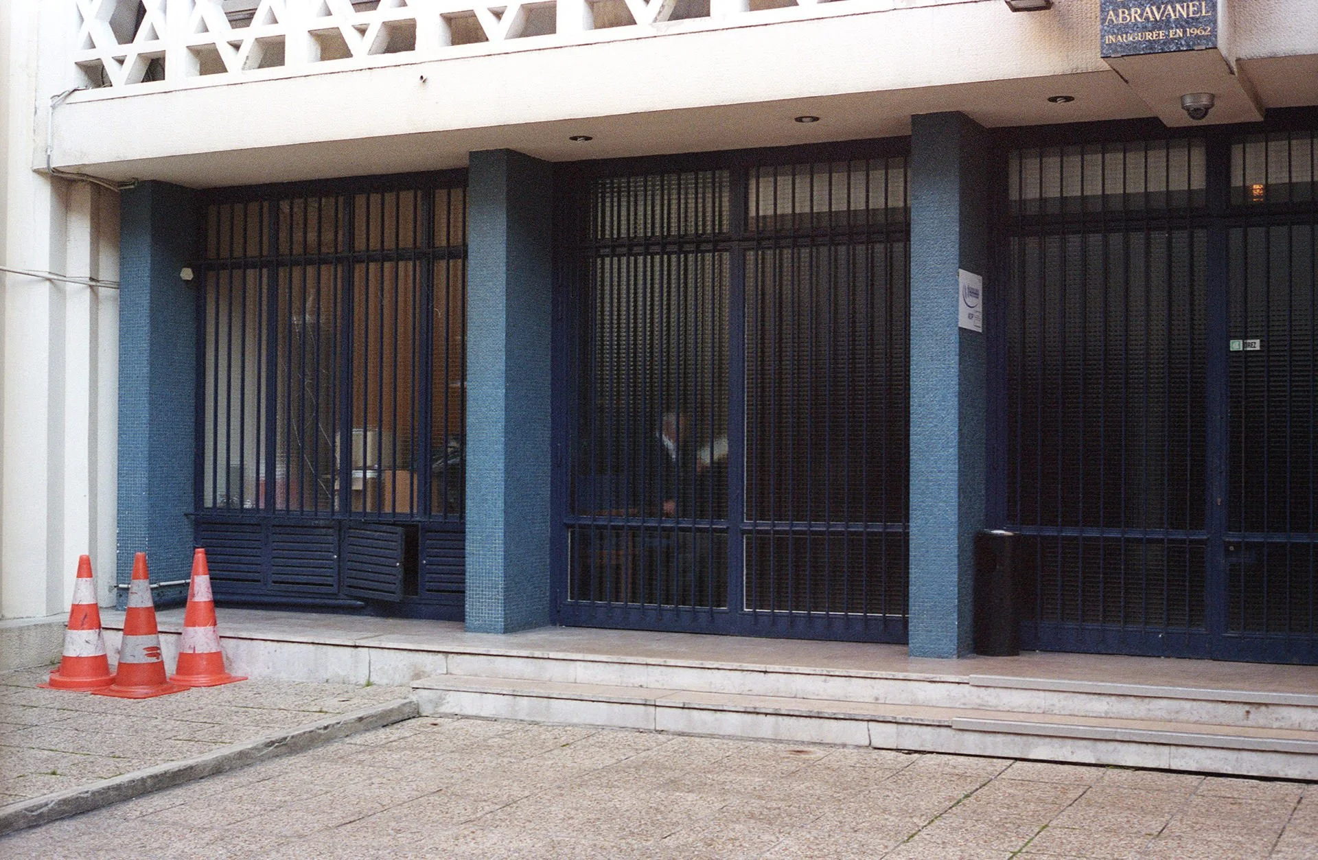 Closed building with black metal gate, blue textured columns, orange traffic cones, and a sidewalk.