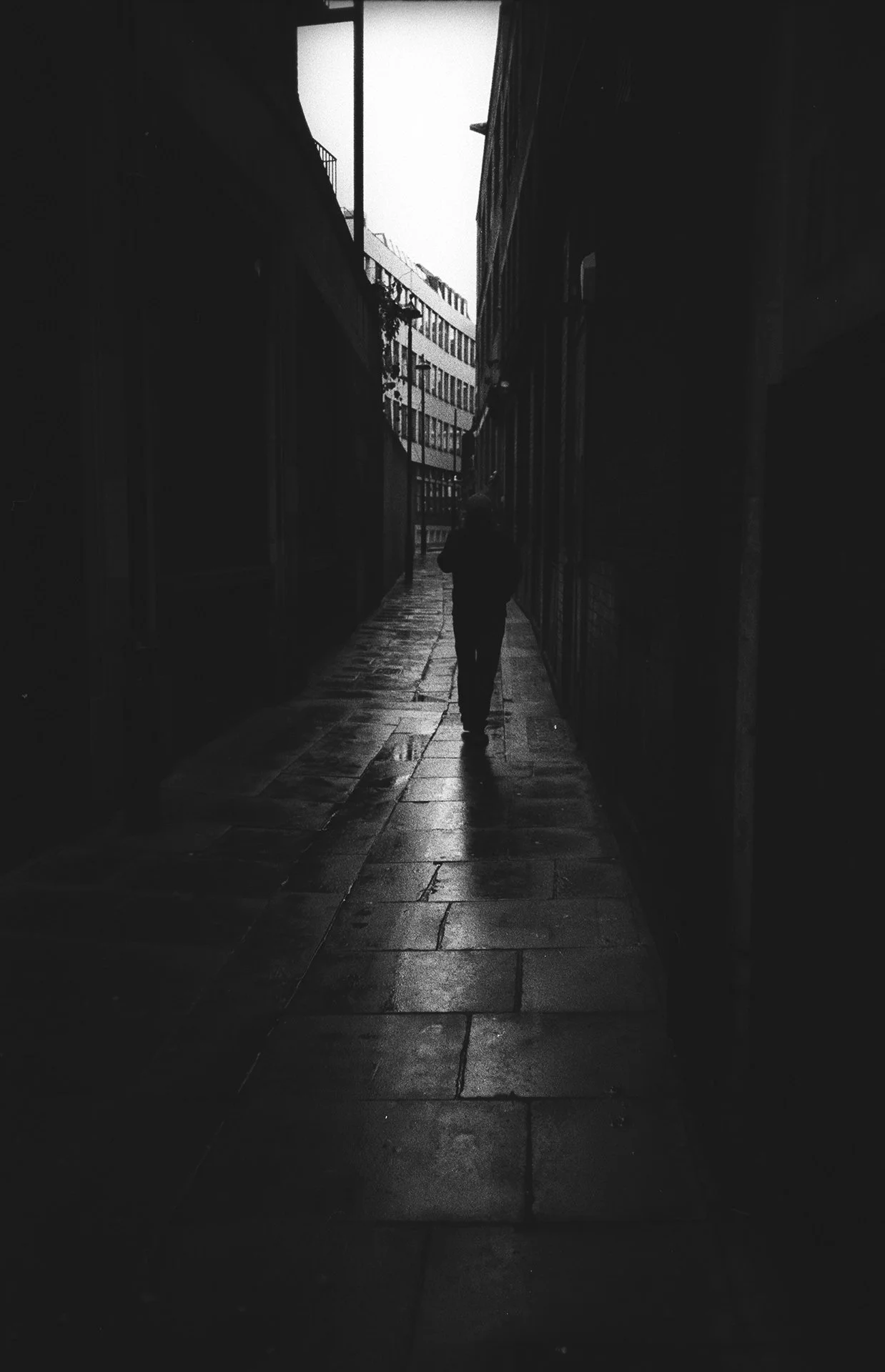 A person walking down a narrow alleyway on a rainy day. The alley is flanked by tall buildings, and the wet pavement reflects light from the sky.
