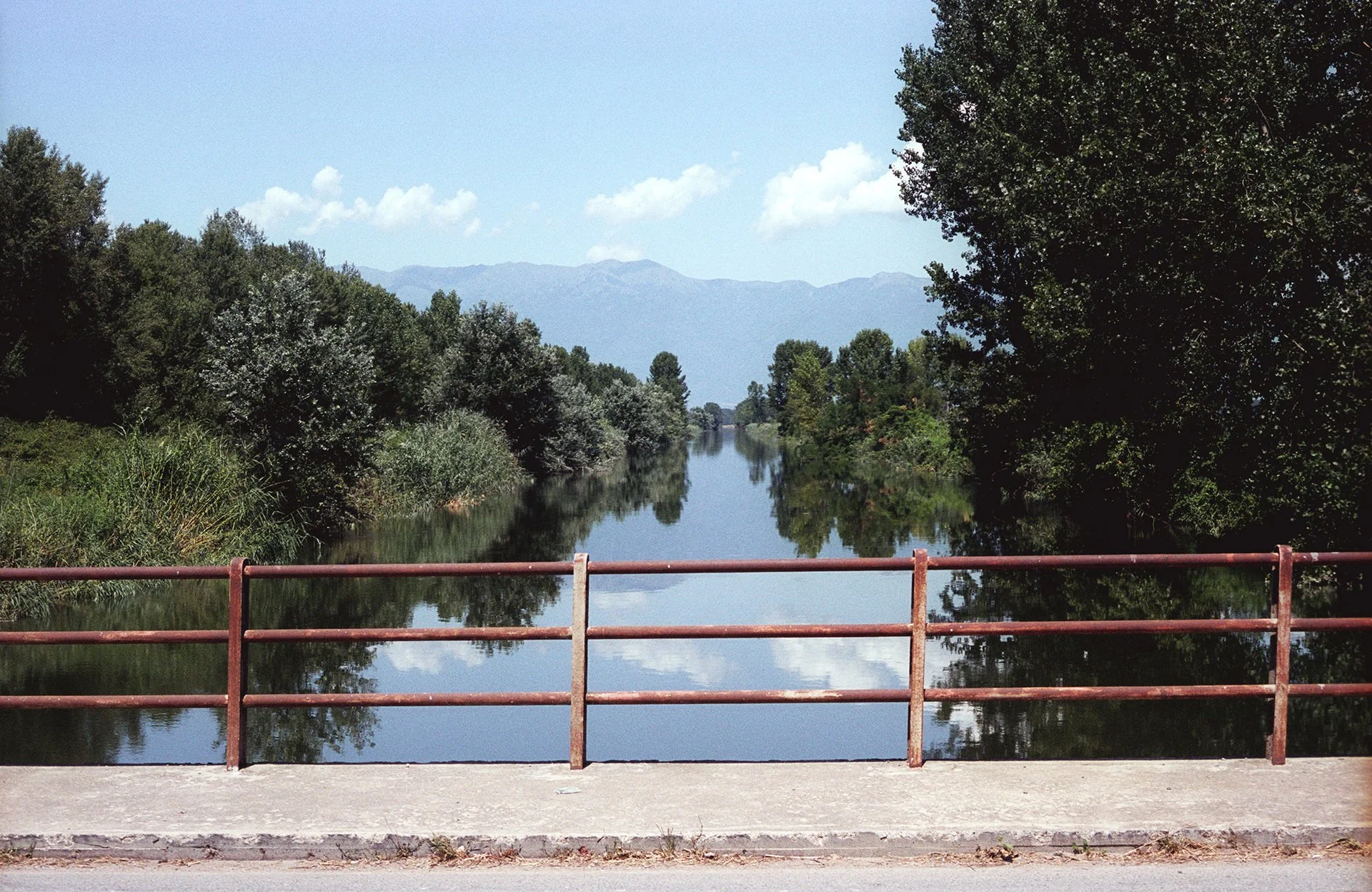 A calm river flowing through a lush green landscape with trees on both sides and mountain ranges in the distance. The photo is taken from behind a rusty metal railing on a bridge.