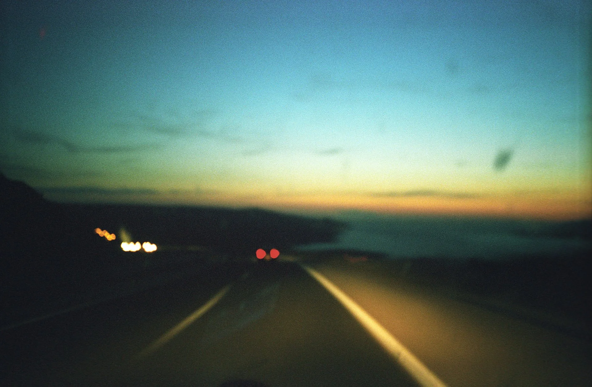 Blurred view of a highway at sunset, with some car lights visible and distant mountains on the horizon.