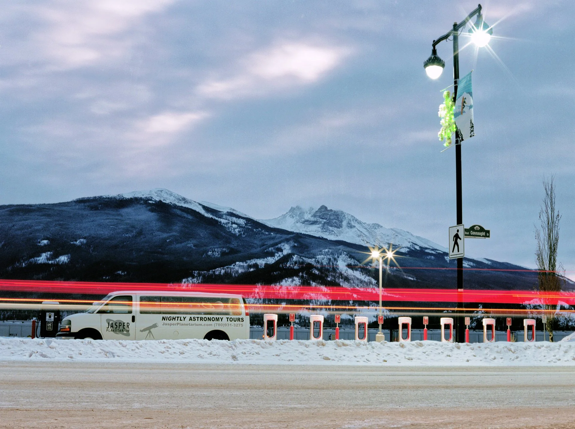 Snow-covered parking lot with a white Jasper Planetarium van, red and white light trails from passing vehicles, streetlights, snow-capped mountains in the background, cloudy sky, and a street sign reading 'Boul Connaught Drive'.