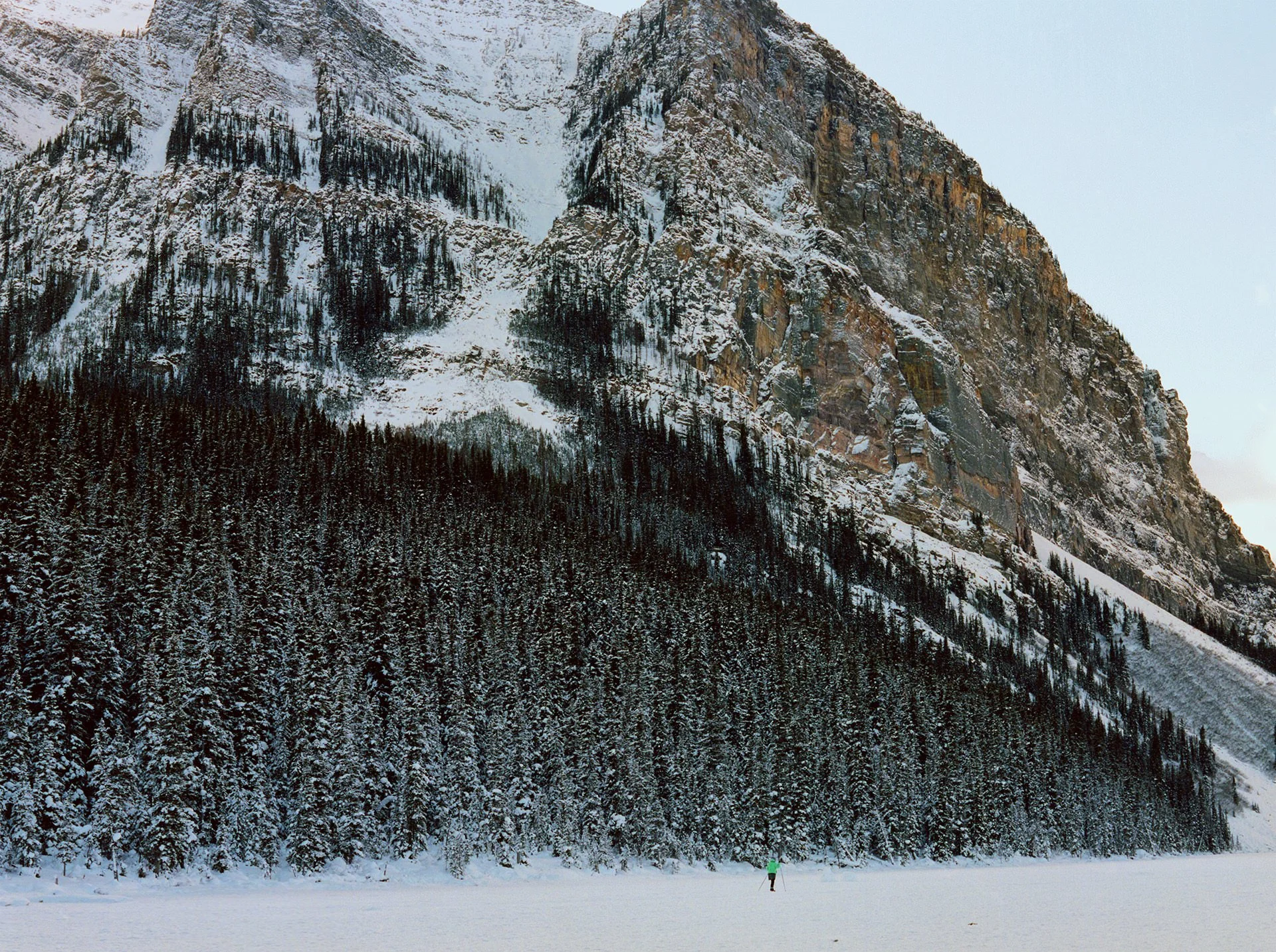 Snow-covered landscape with a mountain and dense forest in the background. A person in a green jacket is walking across a flat, snow-covered ground in the foreground.