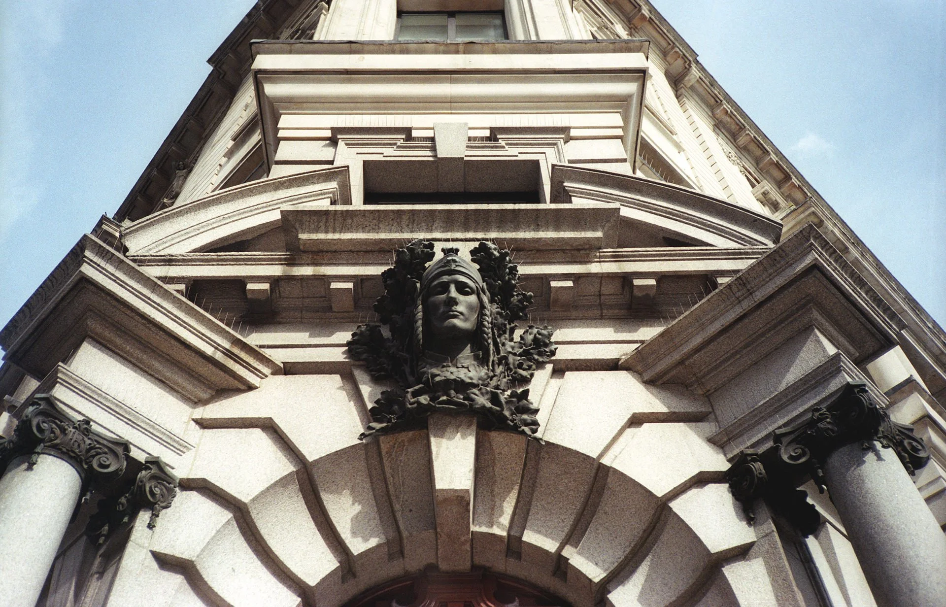 An upward view of an ornate stone building corner with classical architectural details, including a carved female face among floral reliefs, columns with decorative capitals, and a prominent sphere or globe at the top.