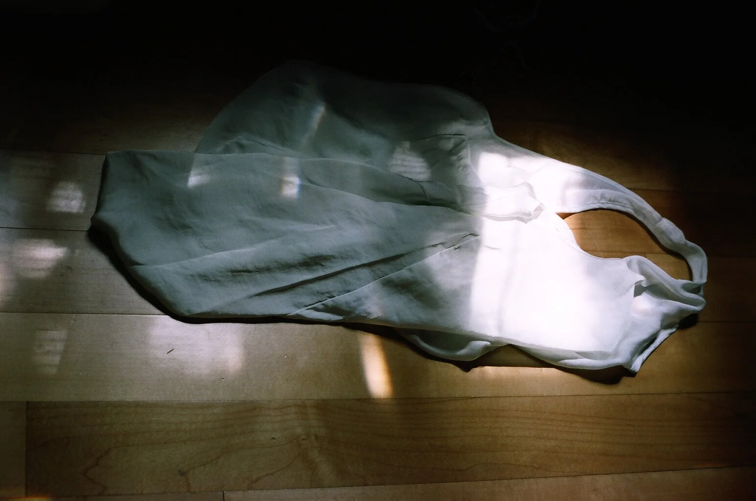 A white silk sleeveless top with a high neck, lying on a wooden floor, illuminated by sunlight.