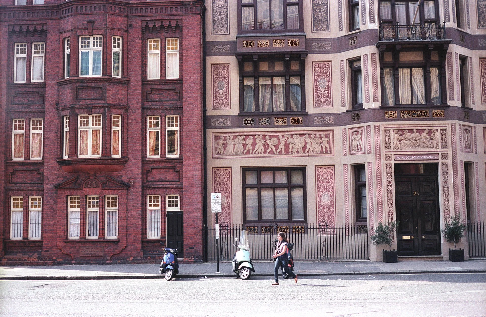 Two ornate buildings, one red brick and one pink with detailed decorative features, on a city street with pedestrians and parked scooters.