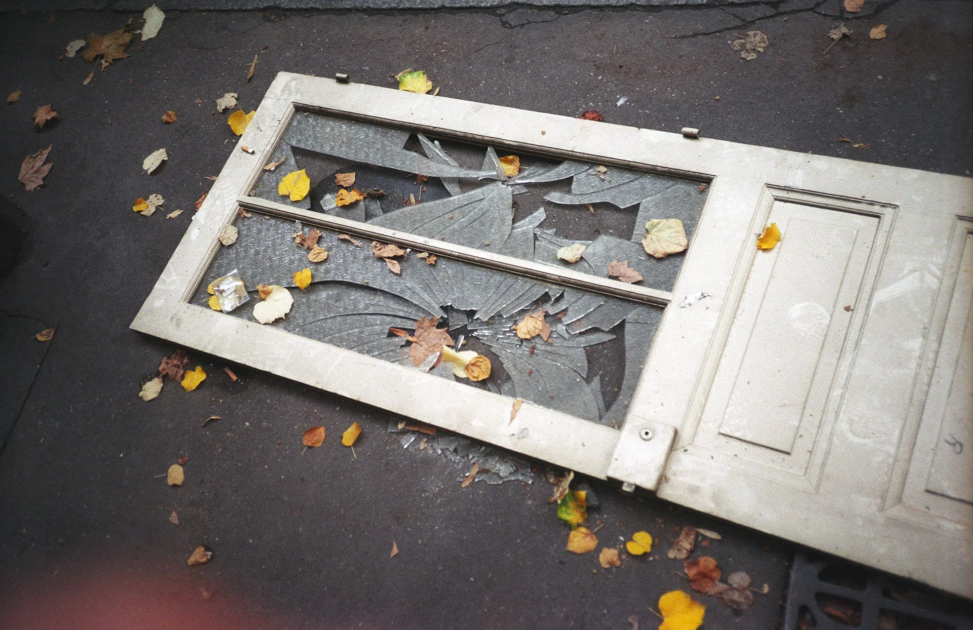 Broken glass door with shattered panels lying on ground surrounded by fallen autumn leaves.