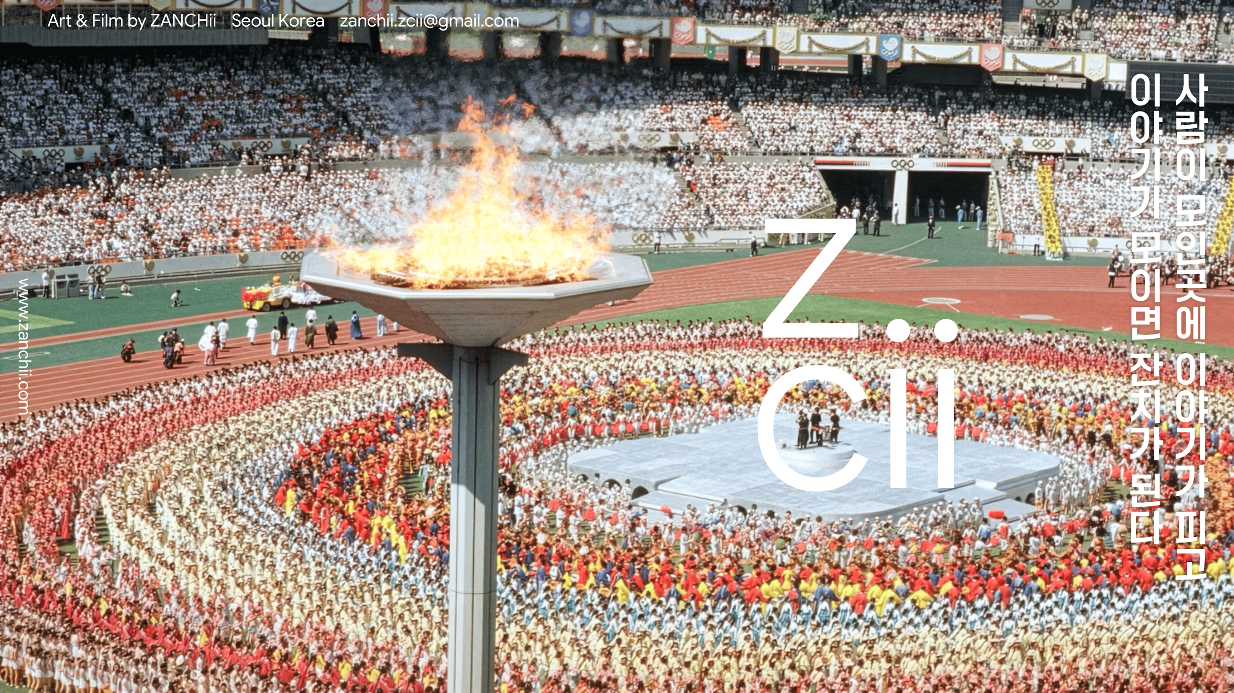 A large outdoor stadium filled with thousands of people, with a fire burning on a raised platform in the center. The crowd appears to be participating in a ceremonial event, with many dressed in white and colorful outfits. The stadium has multiple se