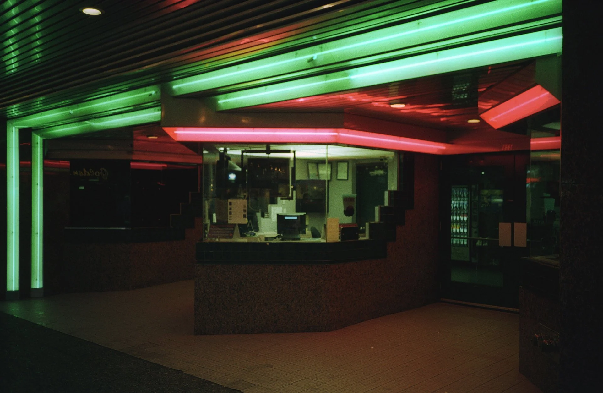 Nighttime view of a fast-food restaurant with neon green and pink lights illuminating the exterior. The restaurant's interior is visible through a glass window, showing a counter and display area.