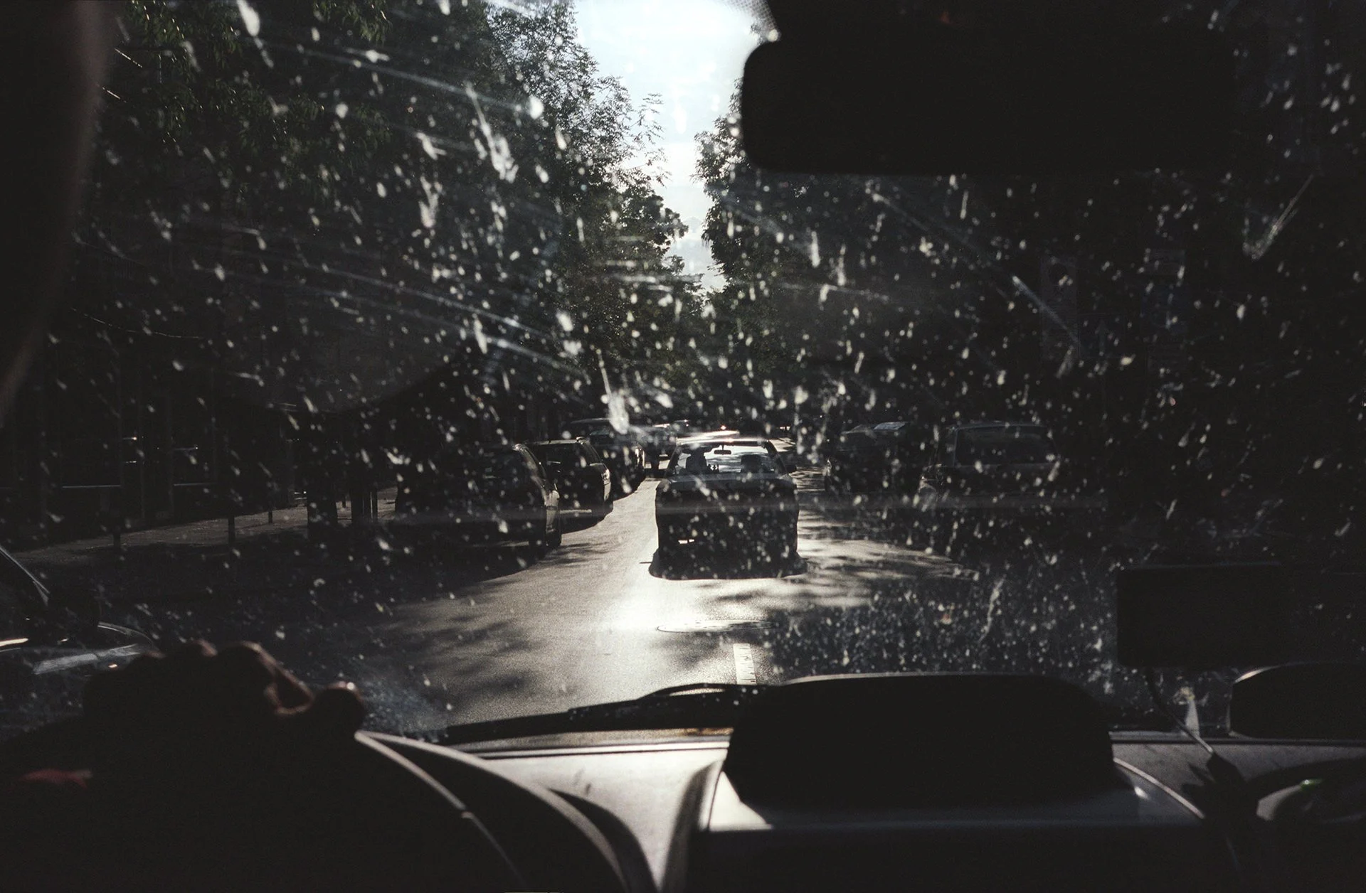 A view from inside a car looking through a rain-covered windshield, showing a street with parked cars and trees, with sunlight reflecting on the wet pavement.