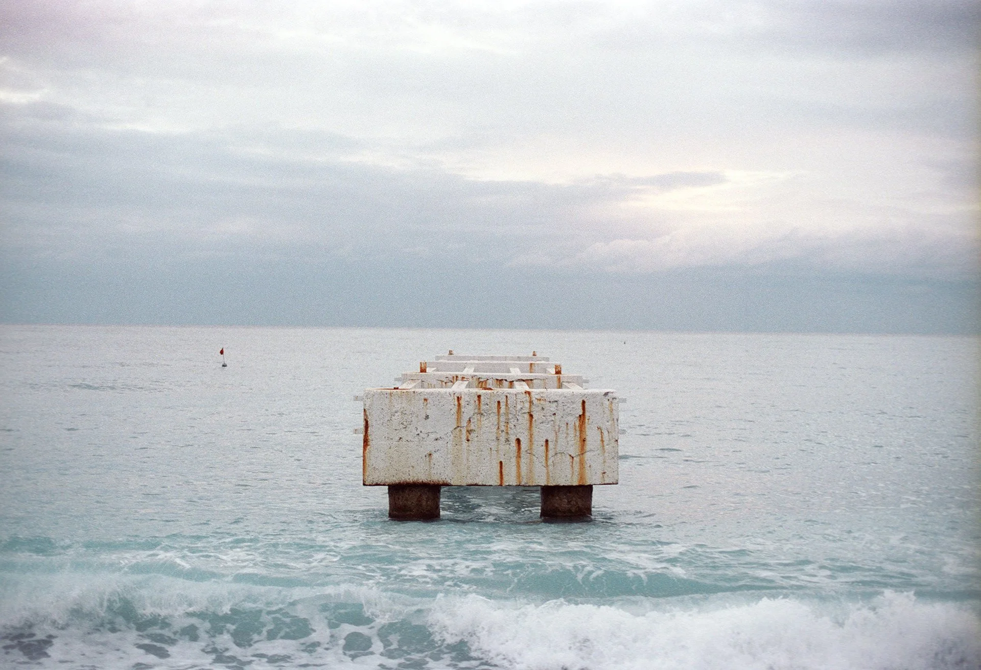A concrete pier extending into the ocean under a cloudy sky, with some rust stains on the pier's surface.