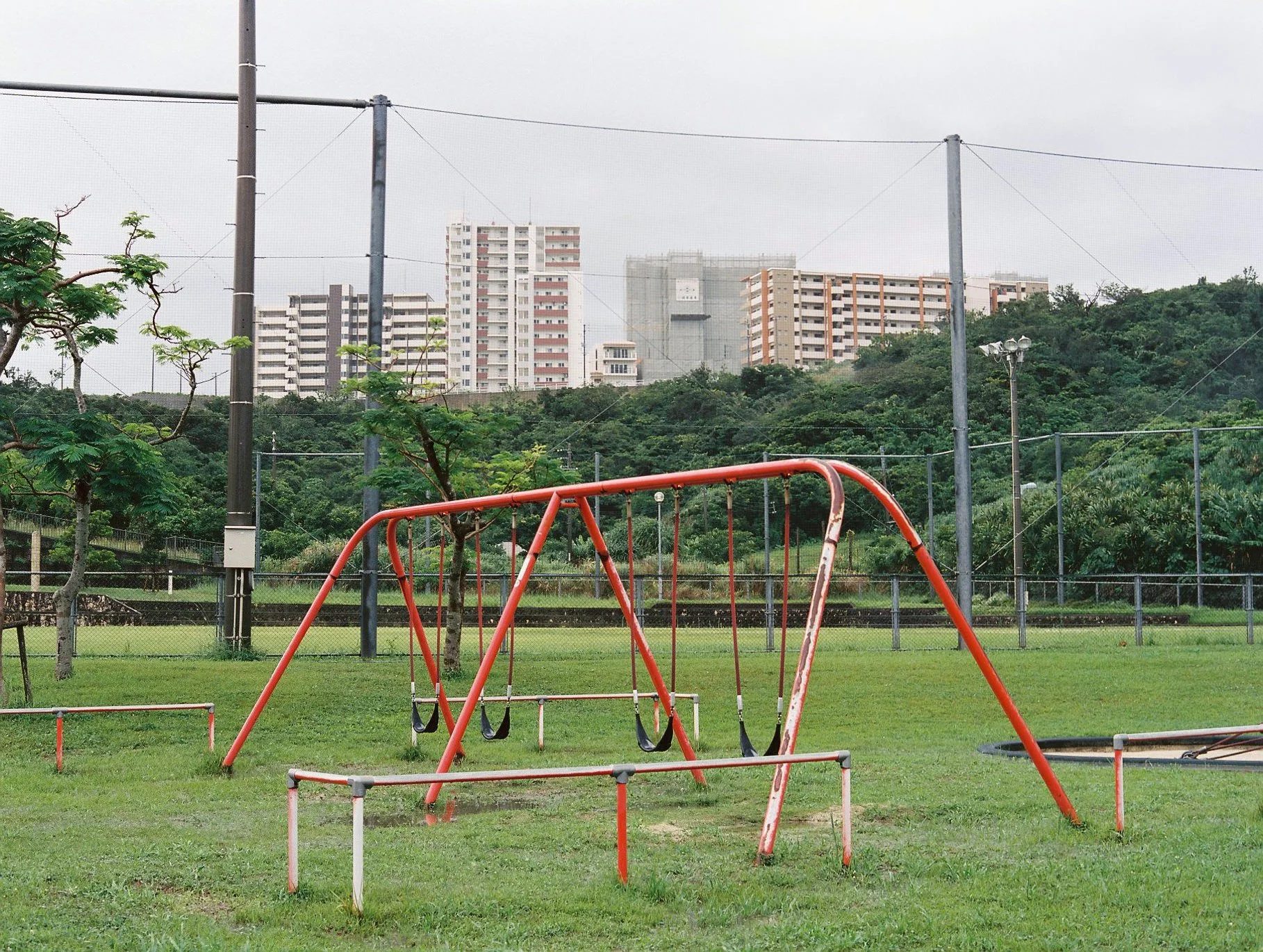 A playground with a red swing set on a grassy field, trees, a chain-link fence, and tall buildings in the background under a cloudy sky.