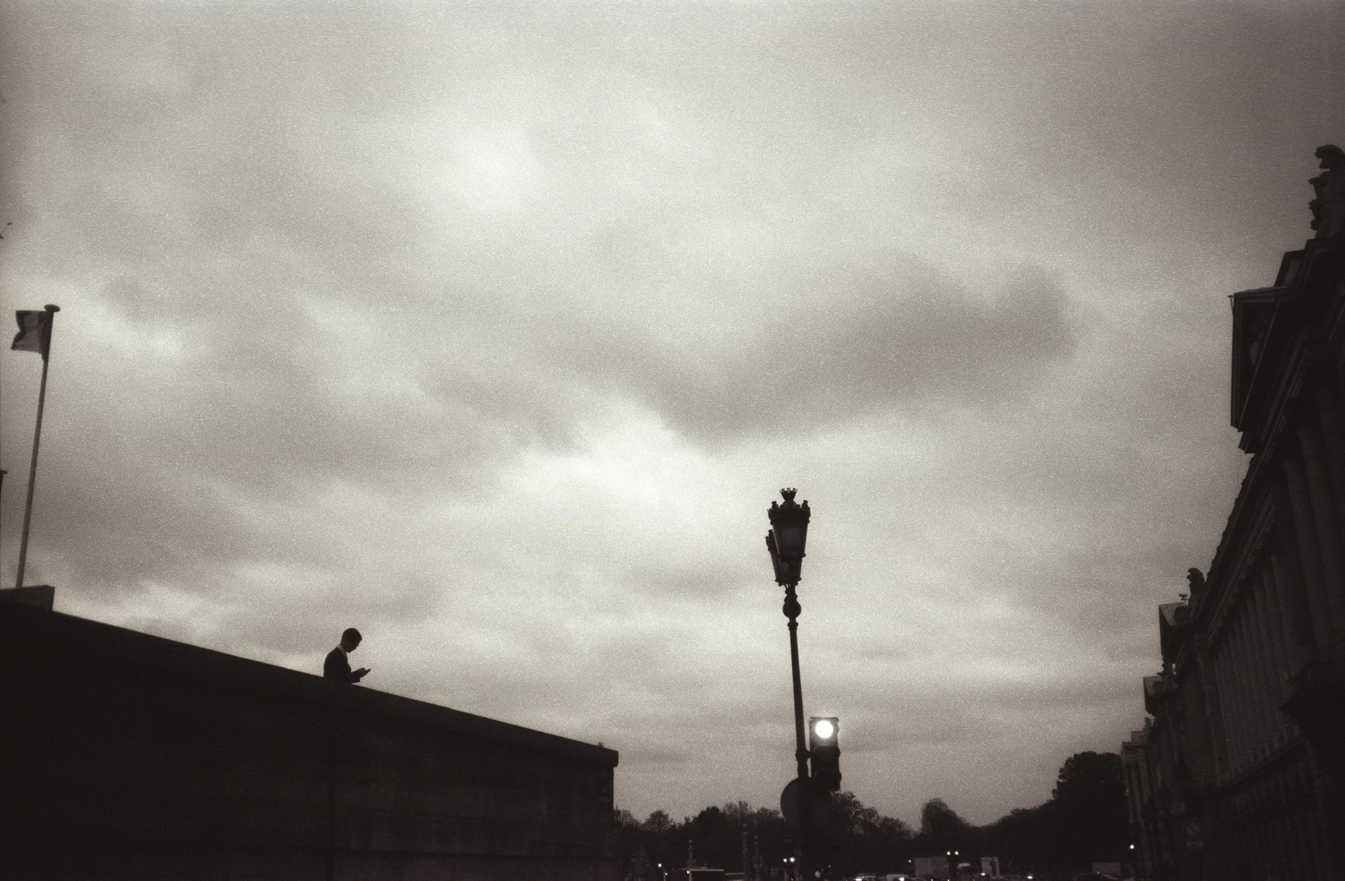 A silhouette of a person looking at their phone, standing on a ledge. The sky is overcast with clouds, and there are streetlights and parts of buildings visible.
