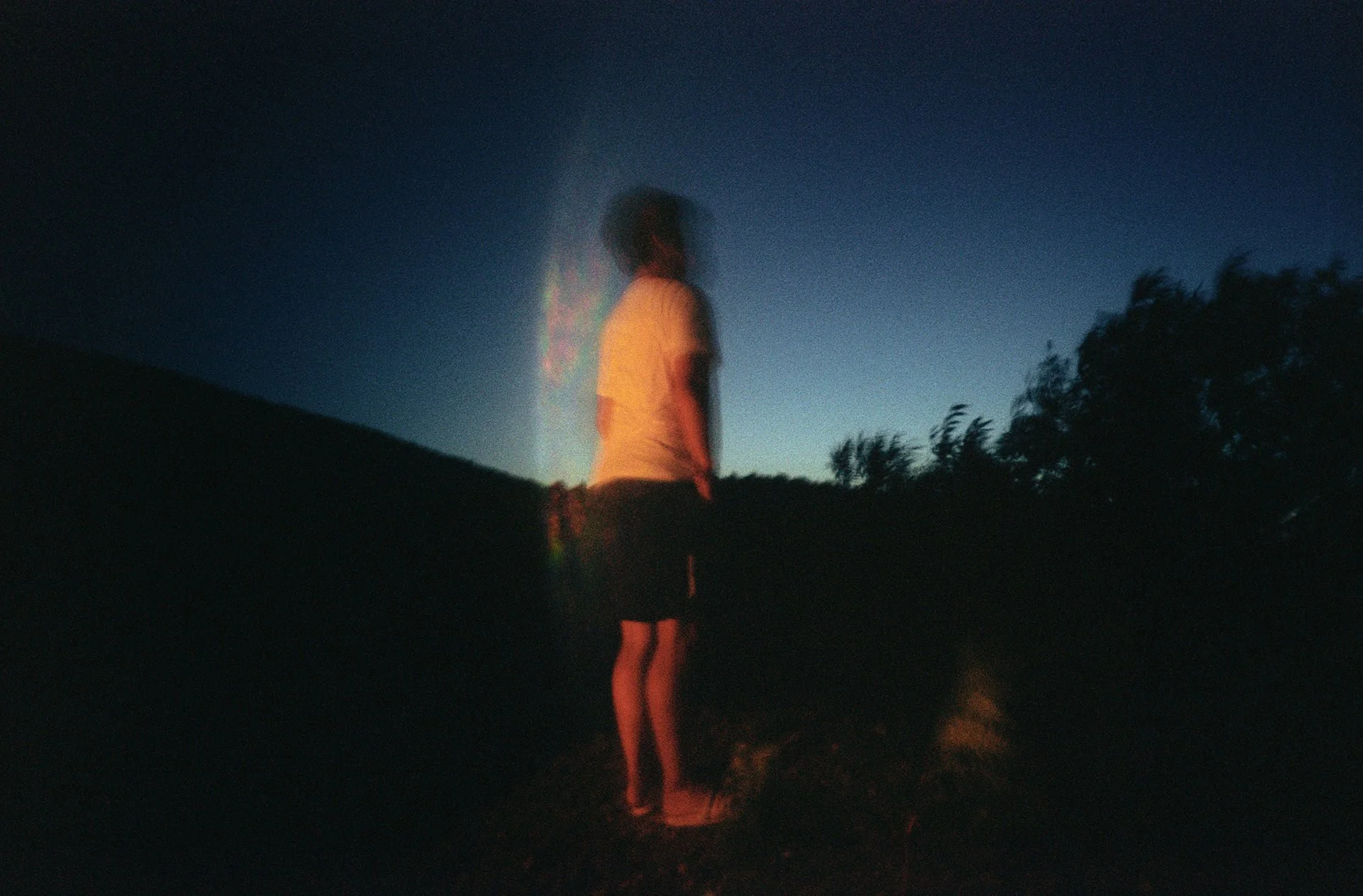 A person standing outdoors at dusk, with a blurred and distorted appearance, with trees in the background and a dark sky.