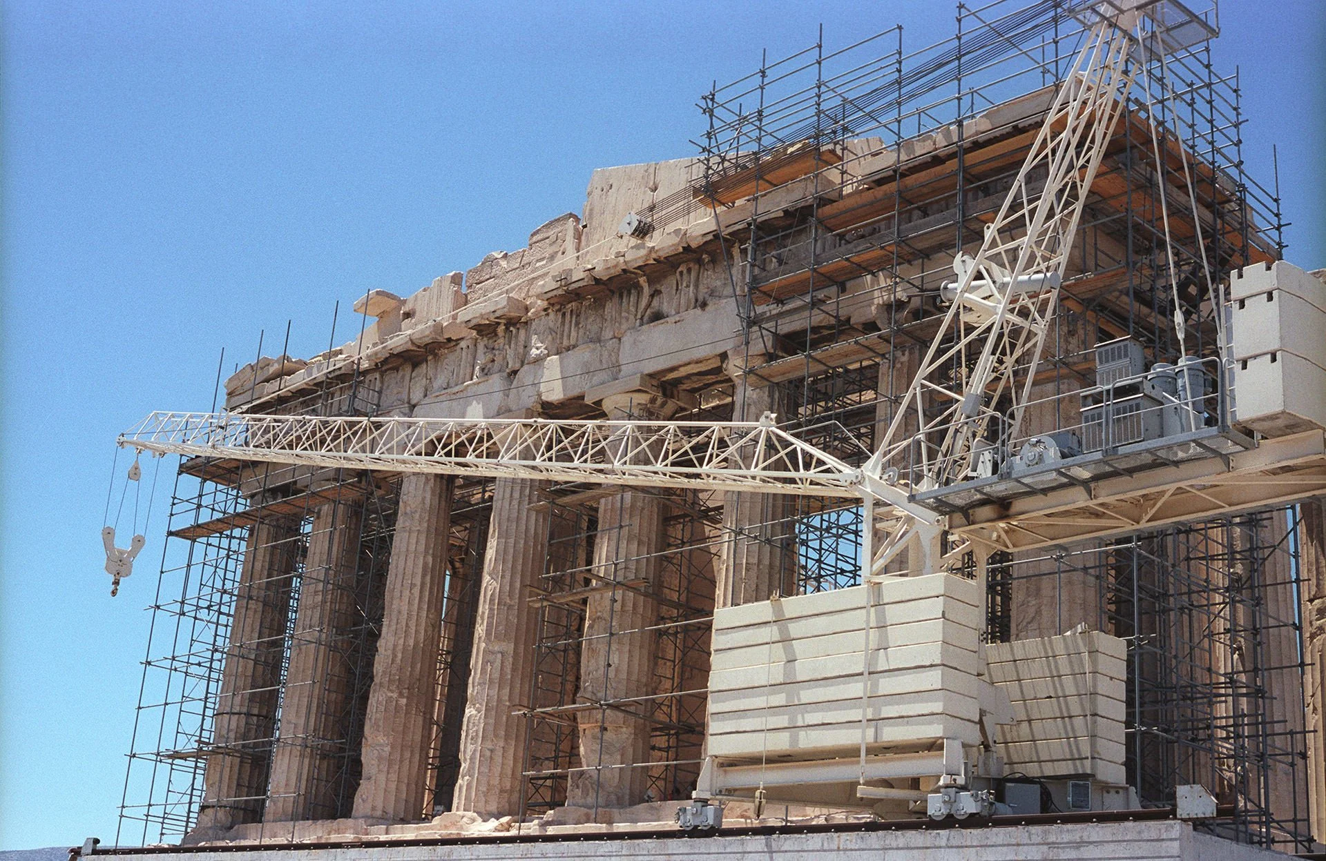 Construction crane beside a partially completed ancient Greek temple with scaffolding, under a clear blue sky.