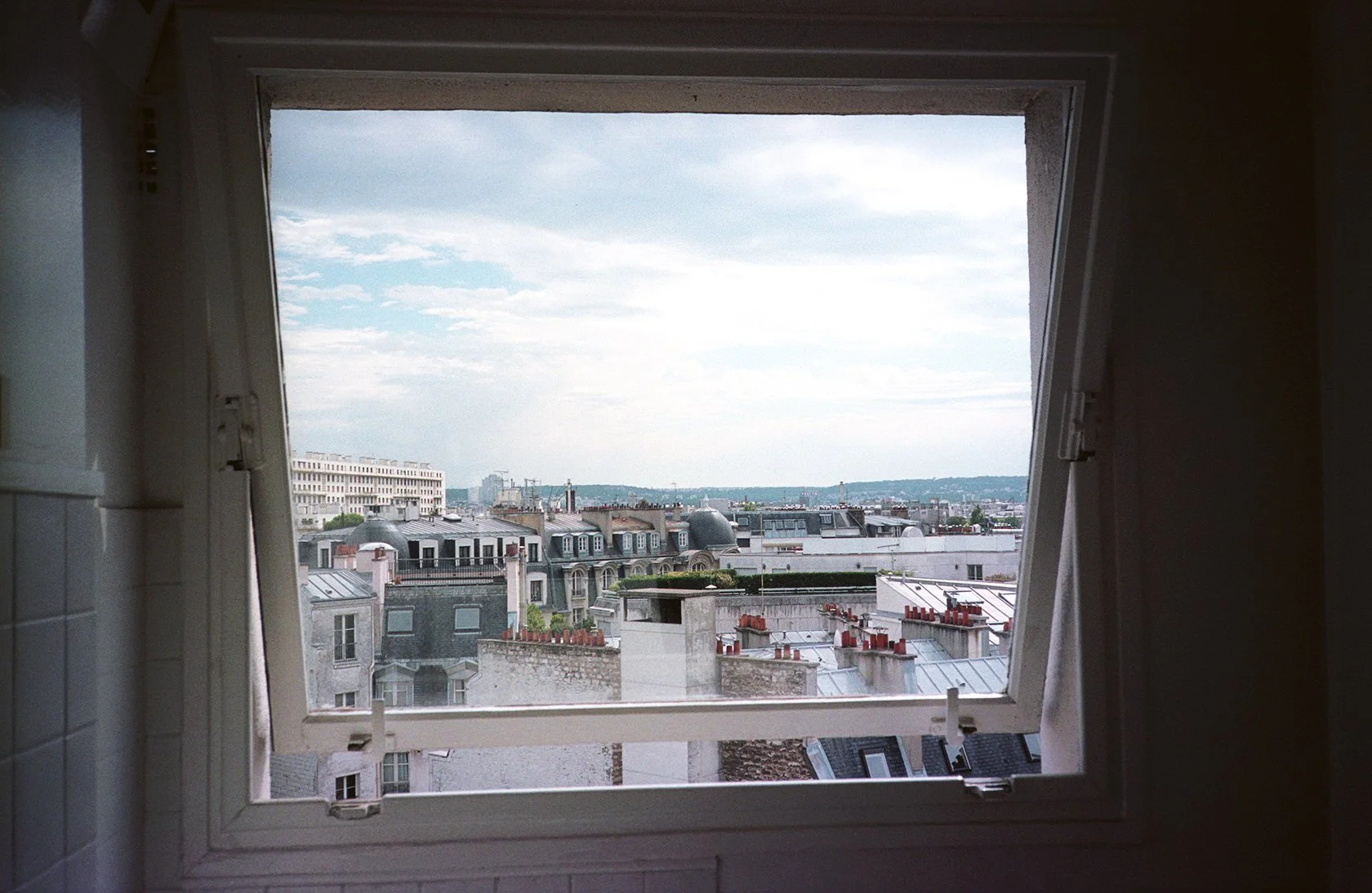 View of a cityscape through a partially open window with white curtains, showing rooftops and buildings with gray and white facades under a cloudy sky.