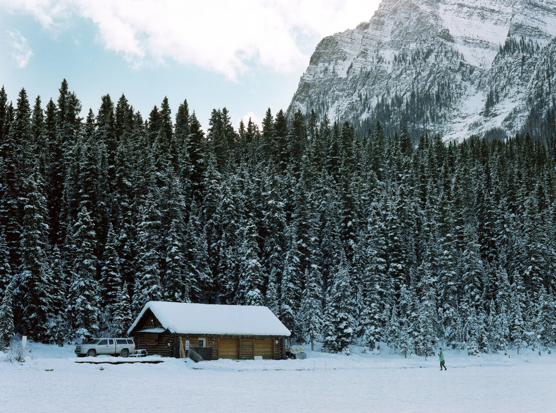 A snow-covered cabin with a car parked in front, surrounded by snowy trees and mountains in the background, with a person walking in the snow.