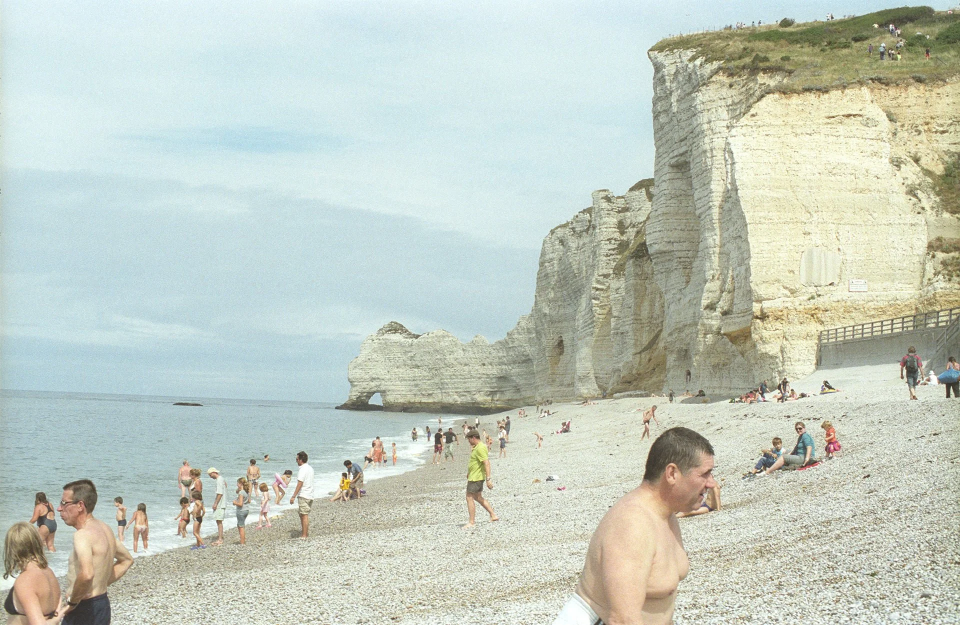 Beach scene with people swimming and sitting on pebble shore, white cliffs with greenery and people at the top in the background, and a cloudy sky.