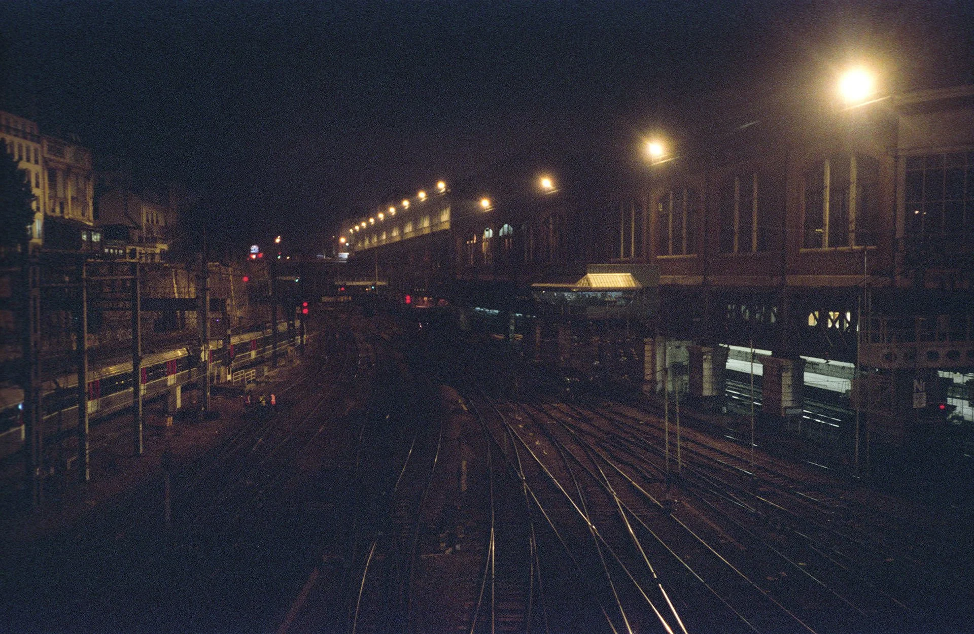 Nighttime view of a city train station with multiple train tracks, urban buildings, and a train on an elevated track, illuminated by streetlights.