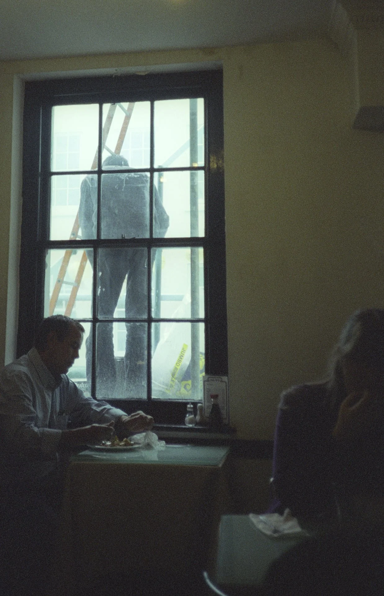 Inside a restaurant, a man is sitting at a table near a window, eating dinner, with another person partially visible in the foreground. Outside the window, a construction worker is visible working on a building.