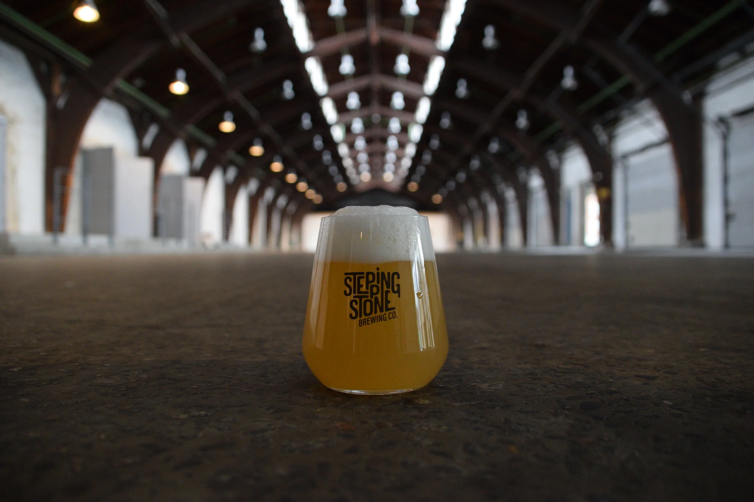 A glass of beer with foam on top, sitting on the ground in an indoor space with a high, arched wooden ceiling and string lights