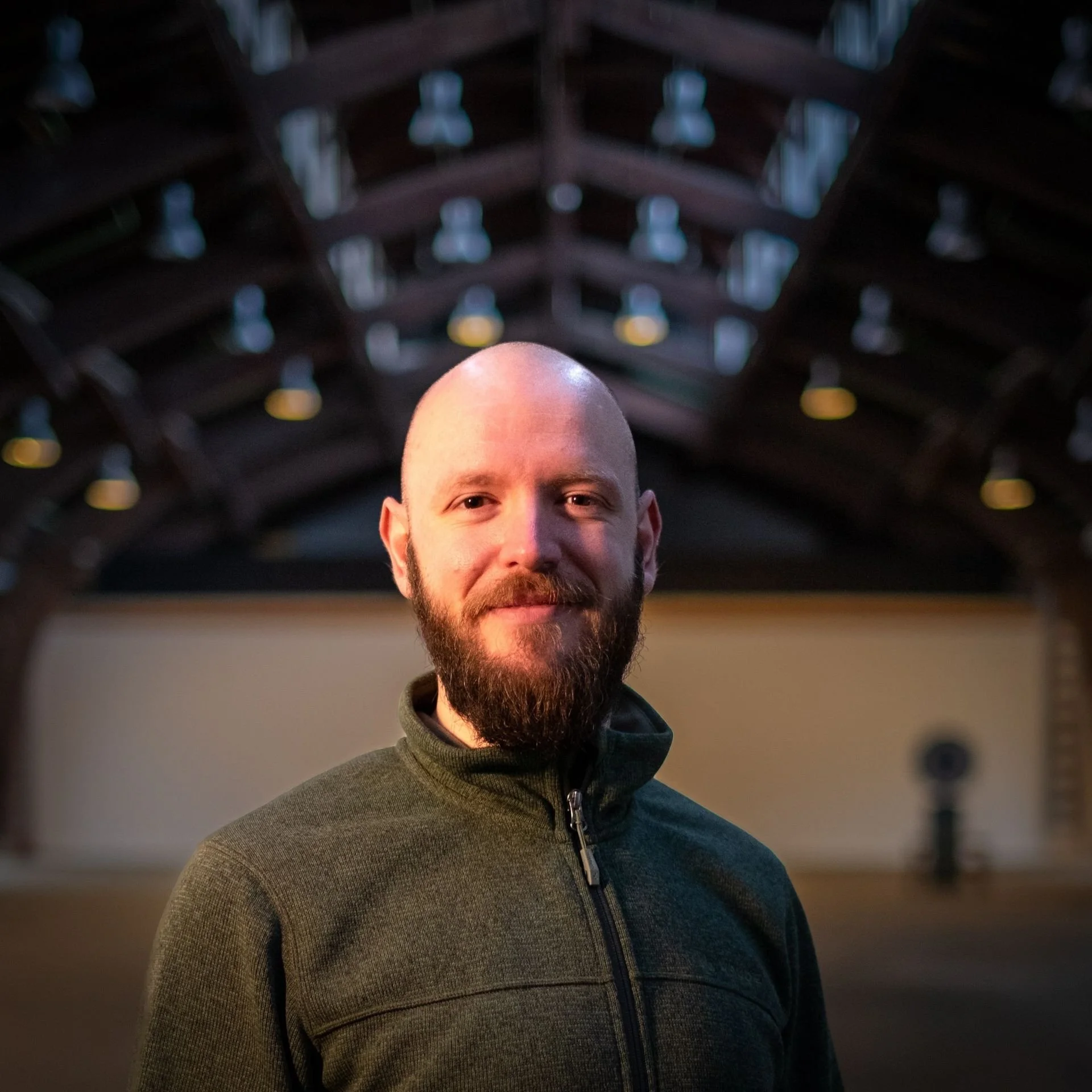 Close-up of a smiling man with a beard and a shaved head, wearing a gray zip-up sweater, standing indoors with a blurred dark, geometric ceiling in the background.