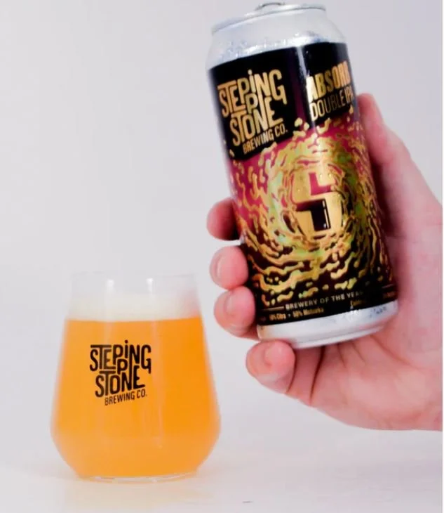 A hand holding a can of Stepping Stone Brewing Co.'s Double IPA beer above a glass filled with the same beer, with the company's logo on both the can and the glass.