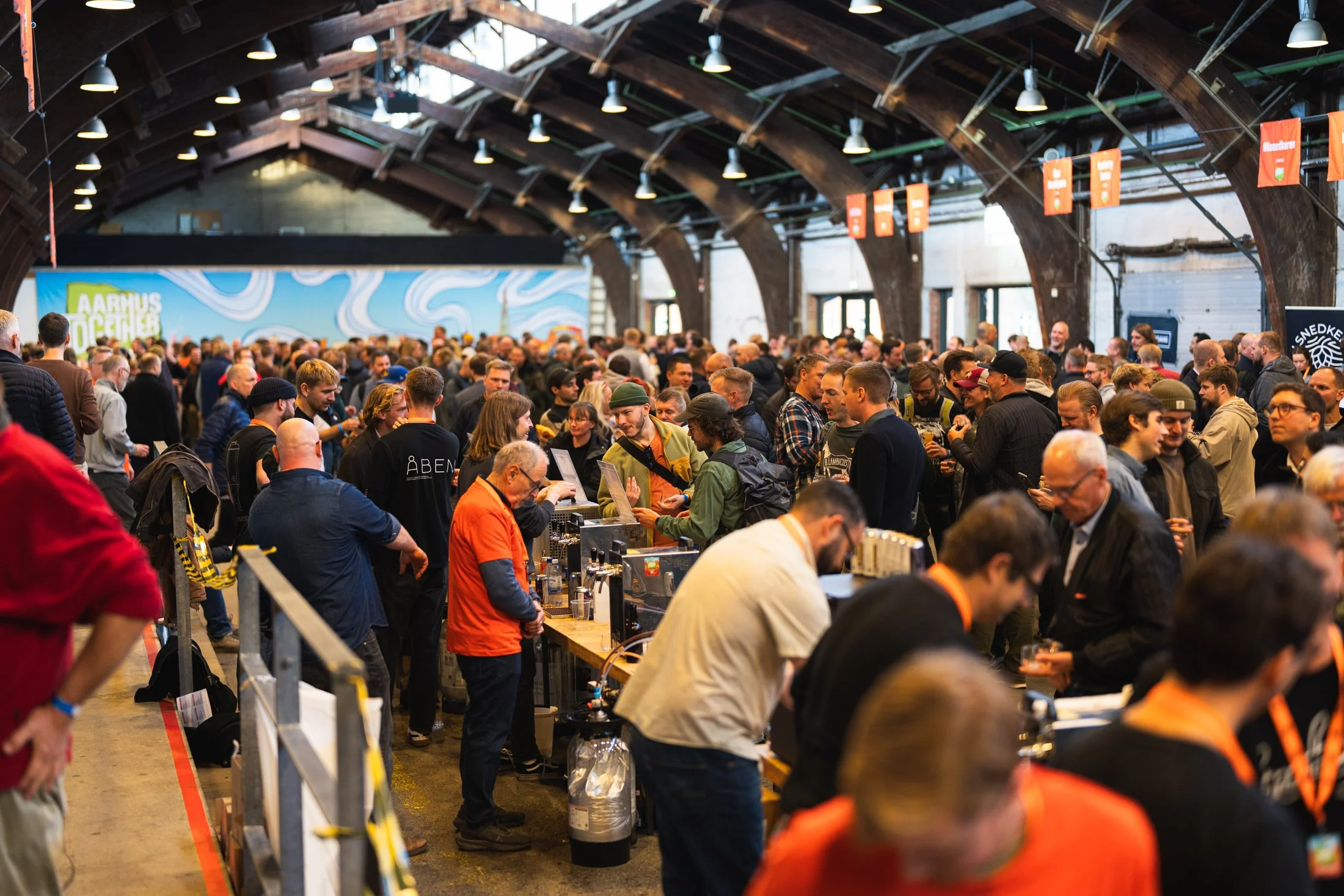 A crowded indoor event with many people gathered around tables and booths, under a high arched wooden ceiling with hanging lights, at the Aarhus Maker Faire.