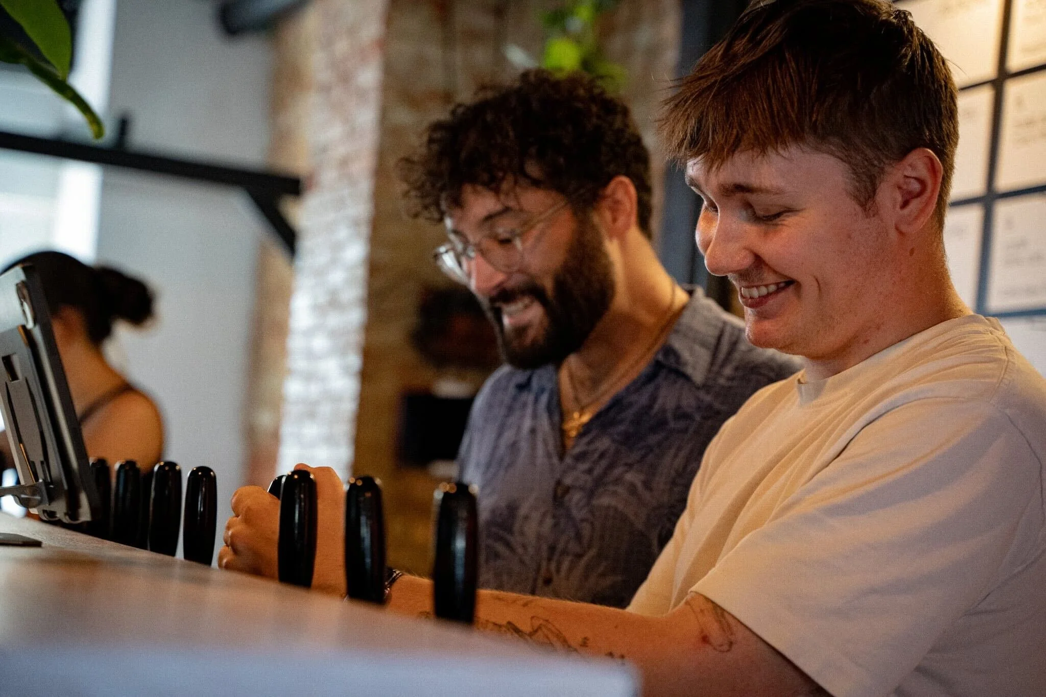 Two men smiling and playing a game with black bottle caps on a wooden counter in a cafe or casual setting, with shelves and a brick wall in the background.