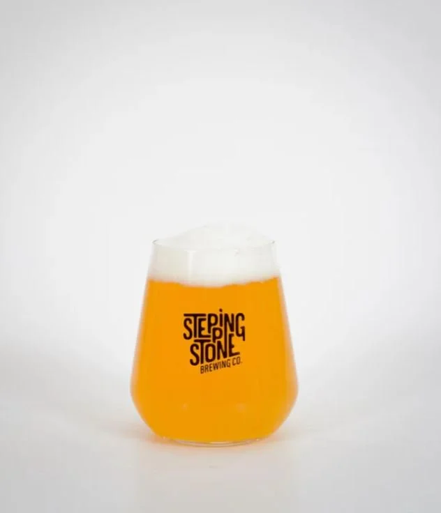 A transparent beer glass with a logo reading 'Stepping Stone Brewing Co.' filled with a hazy orange beer topped with a white foamy head, set against a plain white background.