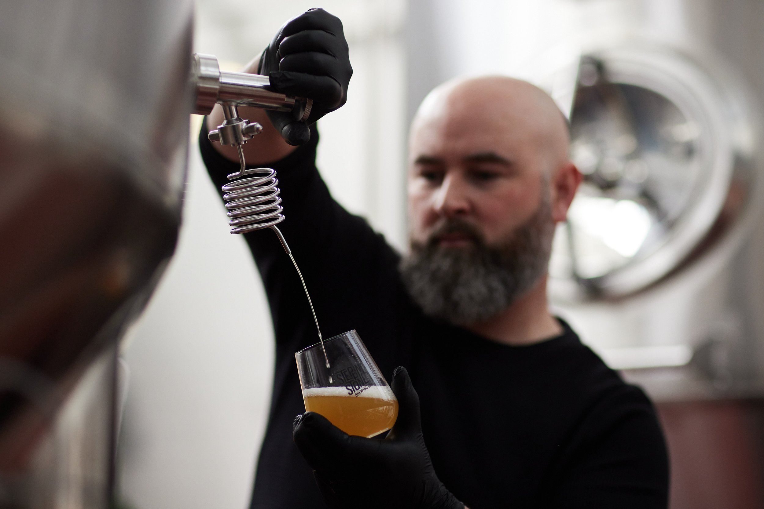 A man with a bald head and beard pouring a golden-colored beverage from a tap into a glass at a brewery.