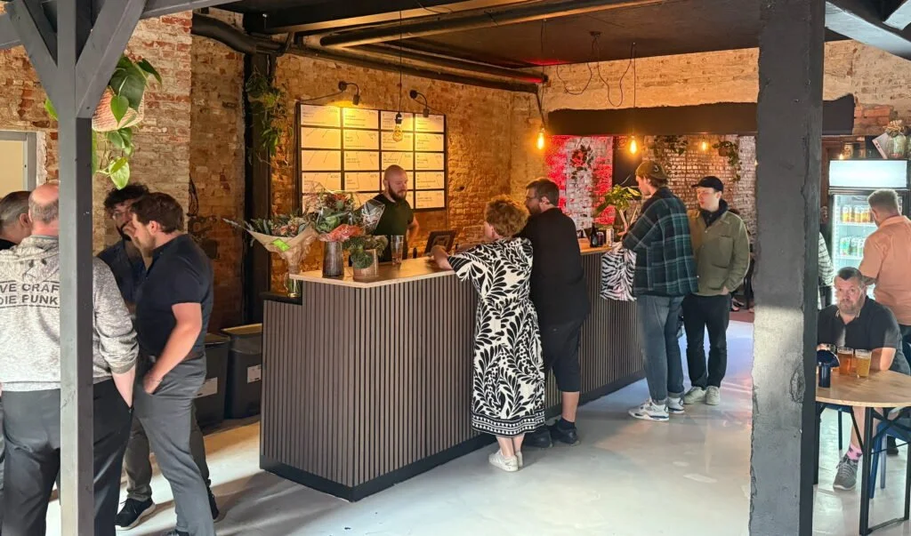 Indtog af bar med flere personer, bartender bag baren, murstensvægge og varm belysning, gæster i baren og nogle der sidder ved borde.