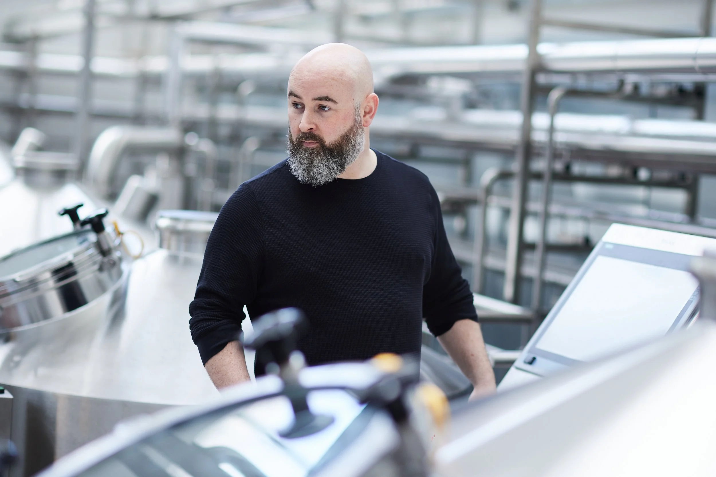A bald man with a beard wearing a black sweater, standing in a metallic industrial or commercial kitchen environment.