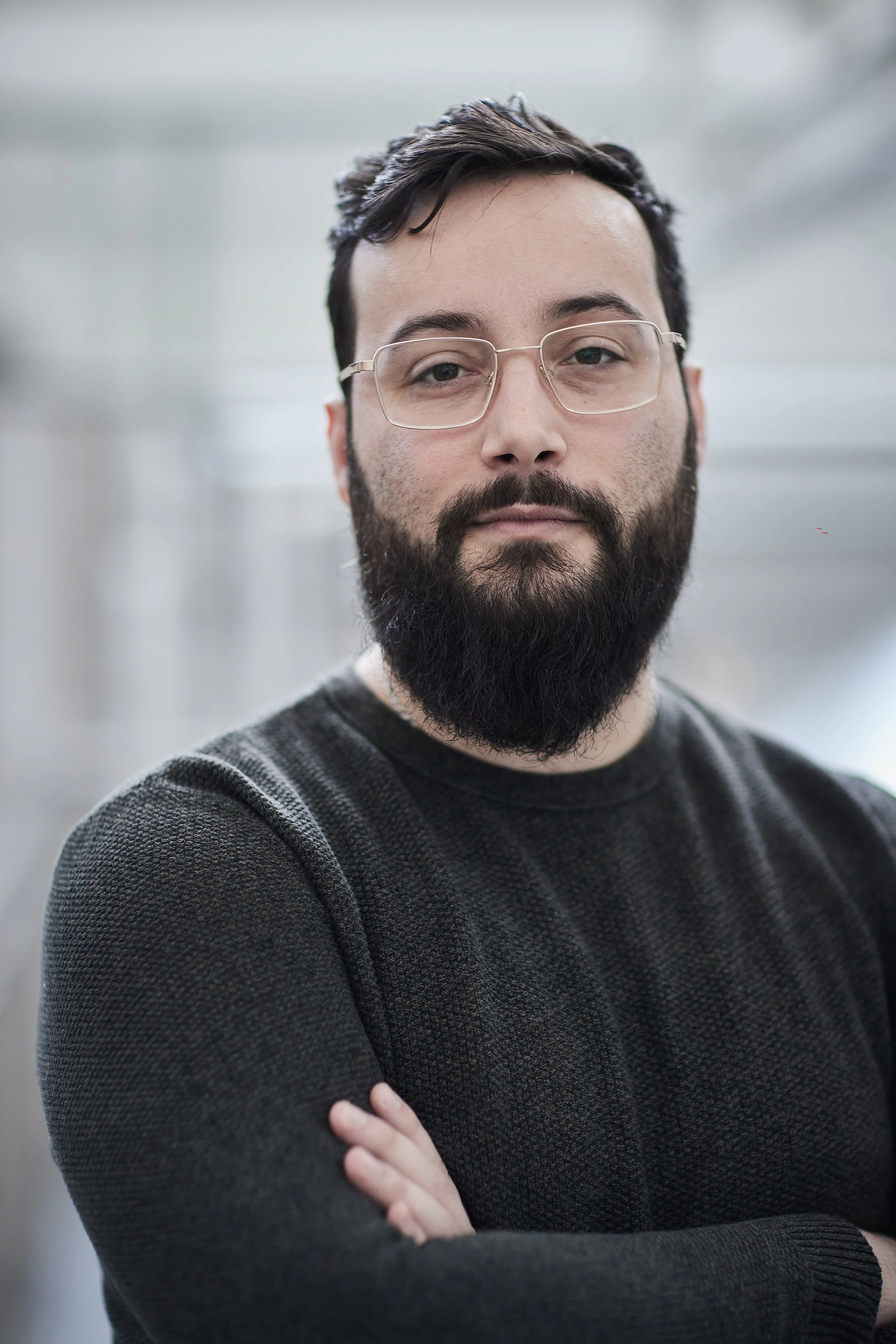 A man with glasses, a beard, and mustache, wearing a gray sweater, standing with crossed arms against a blurred background.