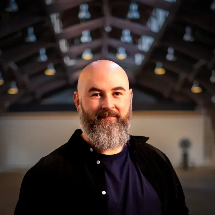 A man with a beard and mustache standing inside a large industrial-style building with exposed wooden beams and hanging lights.