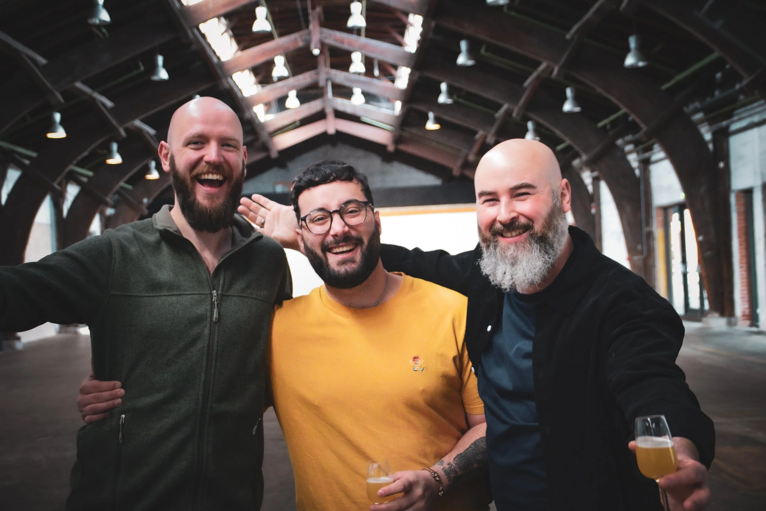 Three men smiling and posing for a photo in an industrial-style venue, with drinks in hand.