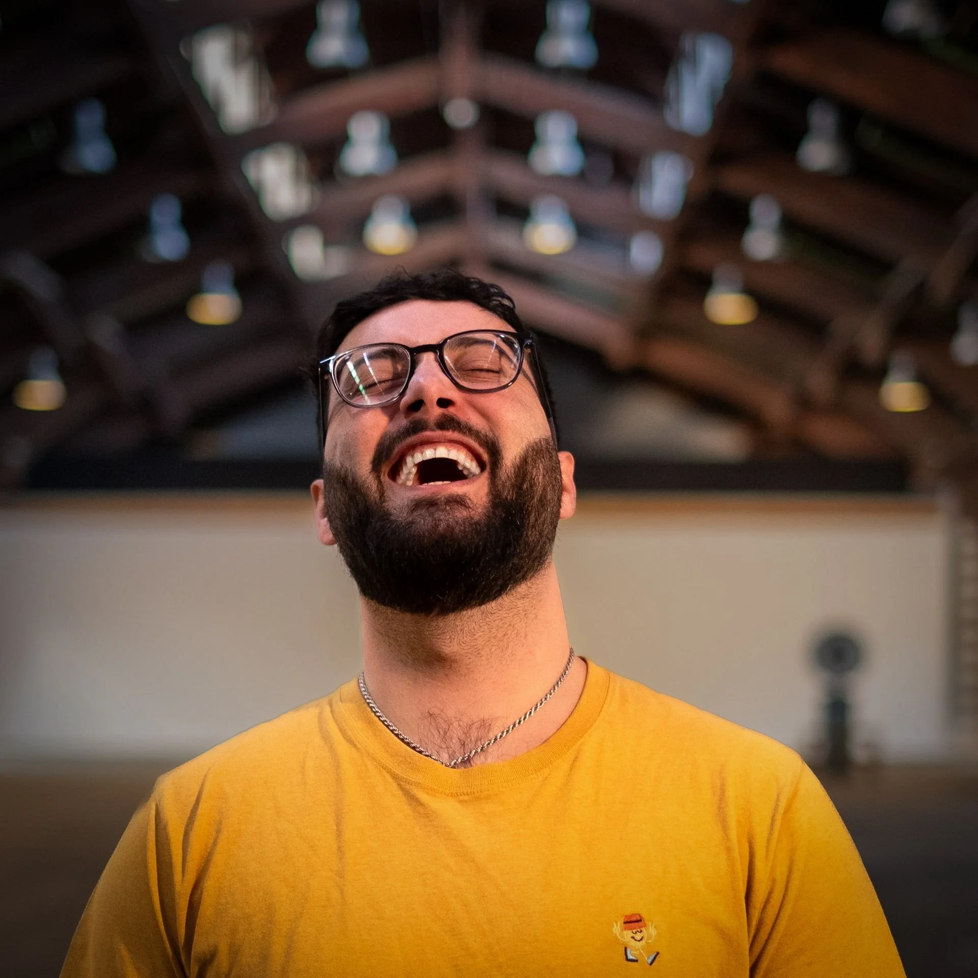 A young man wearing glasses and a yellow t-shirt smiling with his eyes closed, standing indoors with a blurred background.