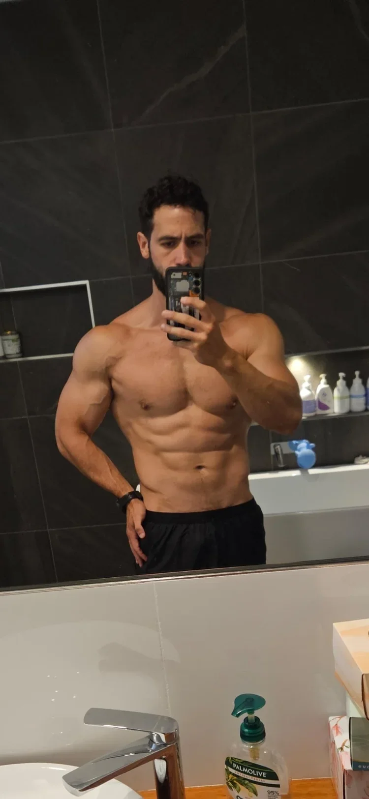 A shirtless man with dark hair and a beard taking a selfie in a bathroom mirror, with dark tiled walls, toiletries on shelves, and a sink with soap in the foreground.
