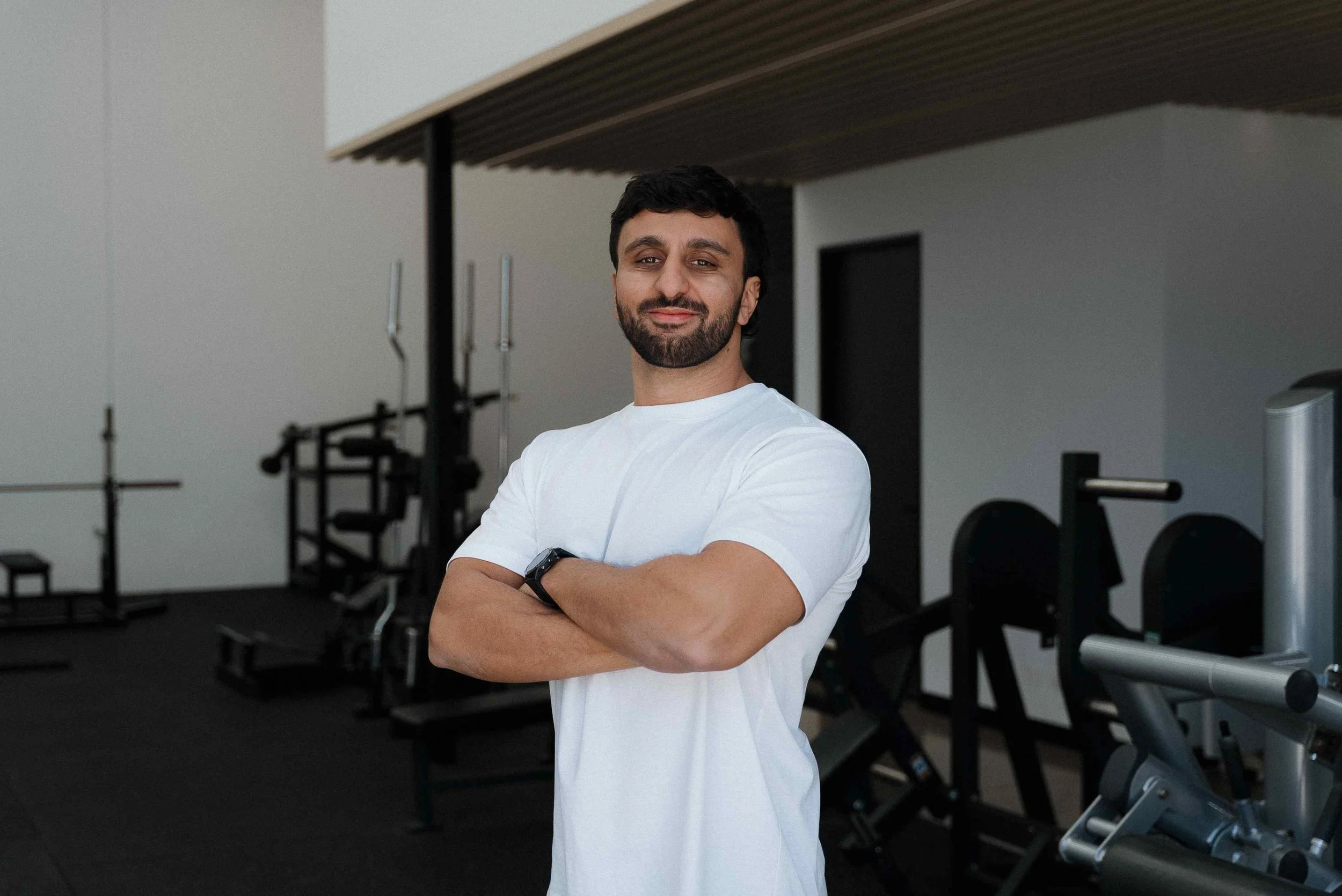 A man with a beard and dark hair standing with arms crossed in a gym or fitness center, smiling slightly, wearing a white T-shirt and a black wristwatch, with gym equipment in the background.