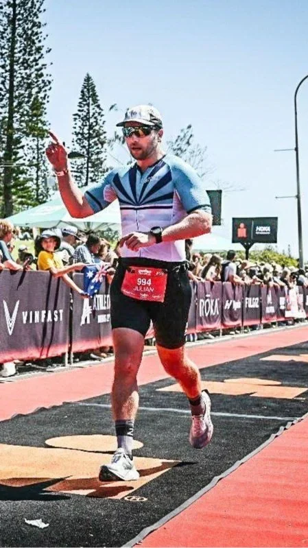 Male marathon runner wearing a race bib, sunglasses, and athletic gear crossing the finish line with spectators behind a barrier in the background.