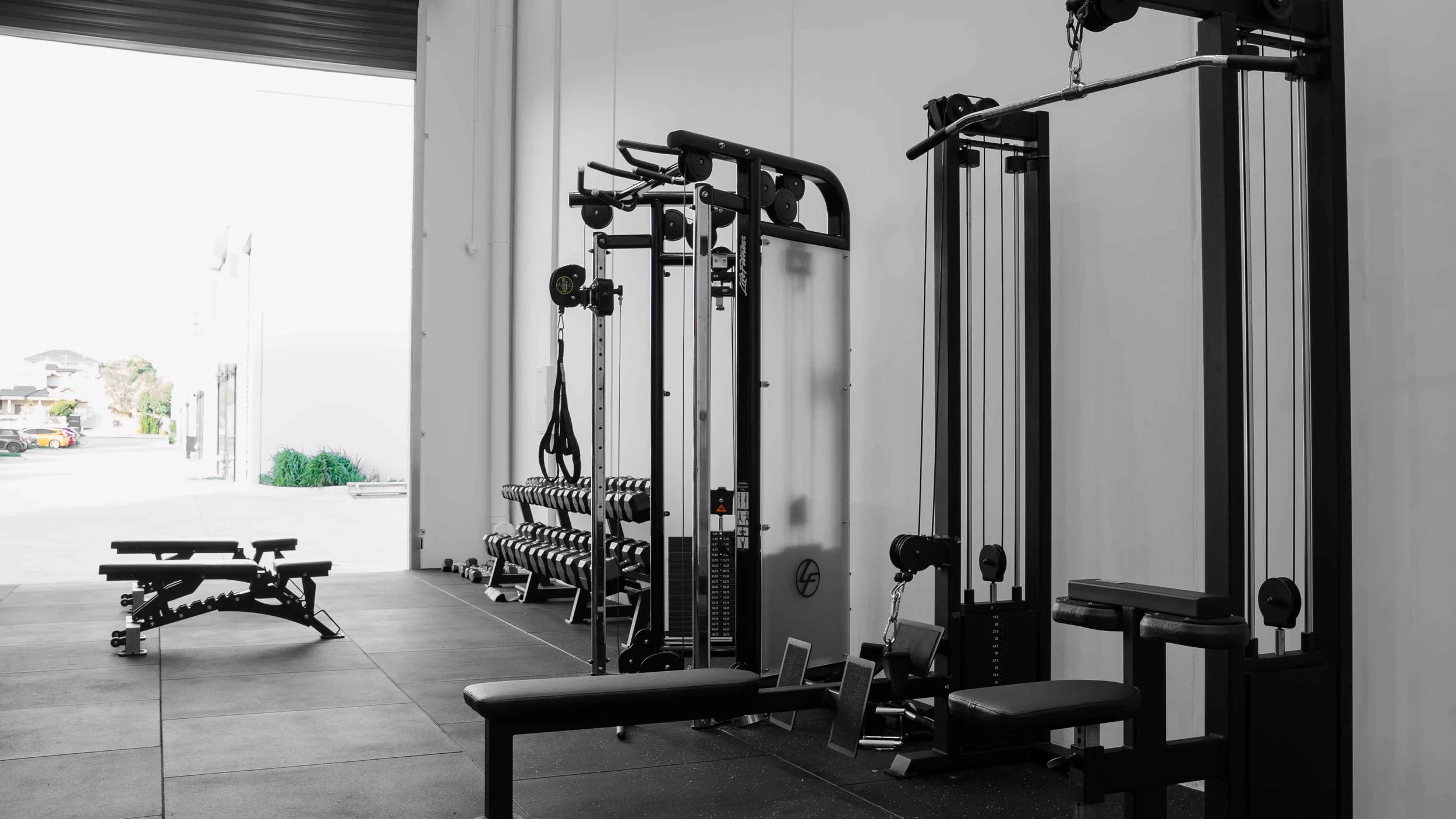 Interior of a gym with various workout equipment including a bench, dumbbells, weight machines, and exercise mats, with large windows showing an outdoor view.