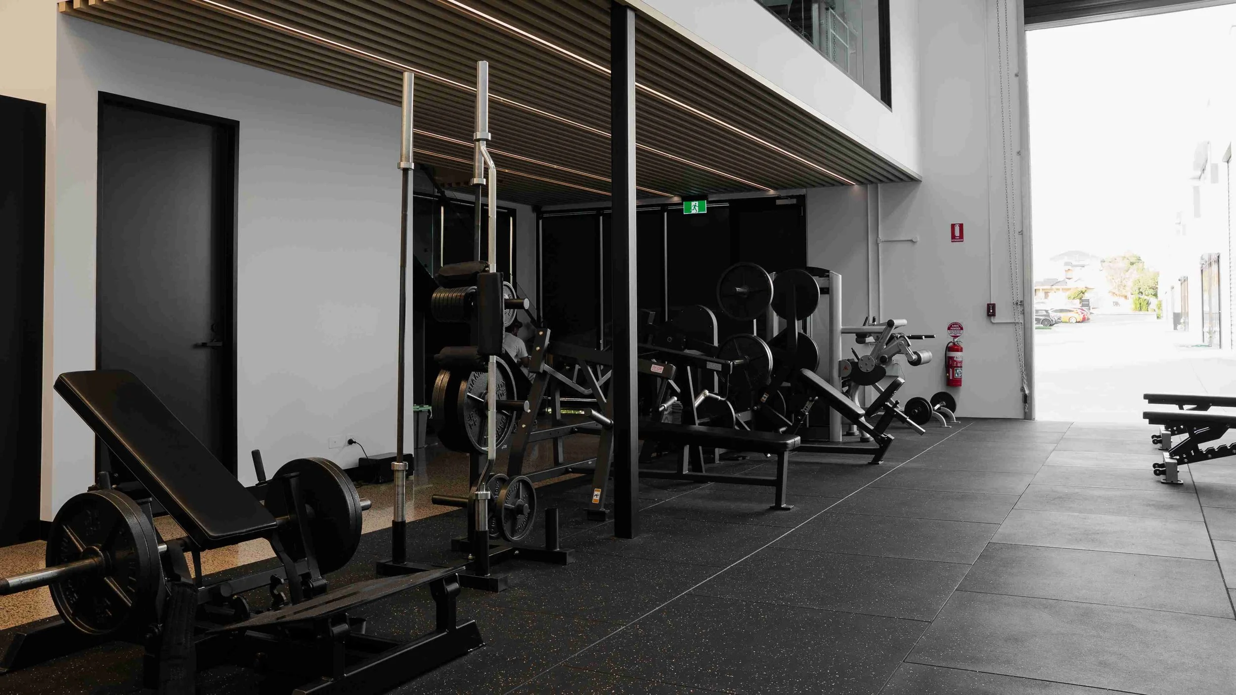 Interior view of a modern gym with various weightlifting and exercise equipment, including dumbbells, benches, and weight machines, with large windows allowing natural light.