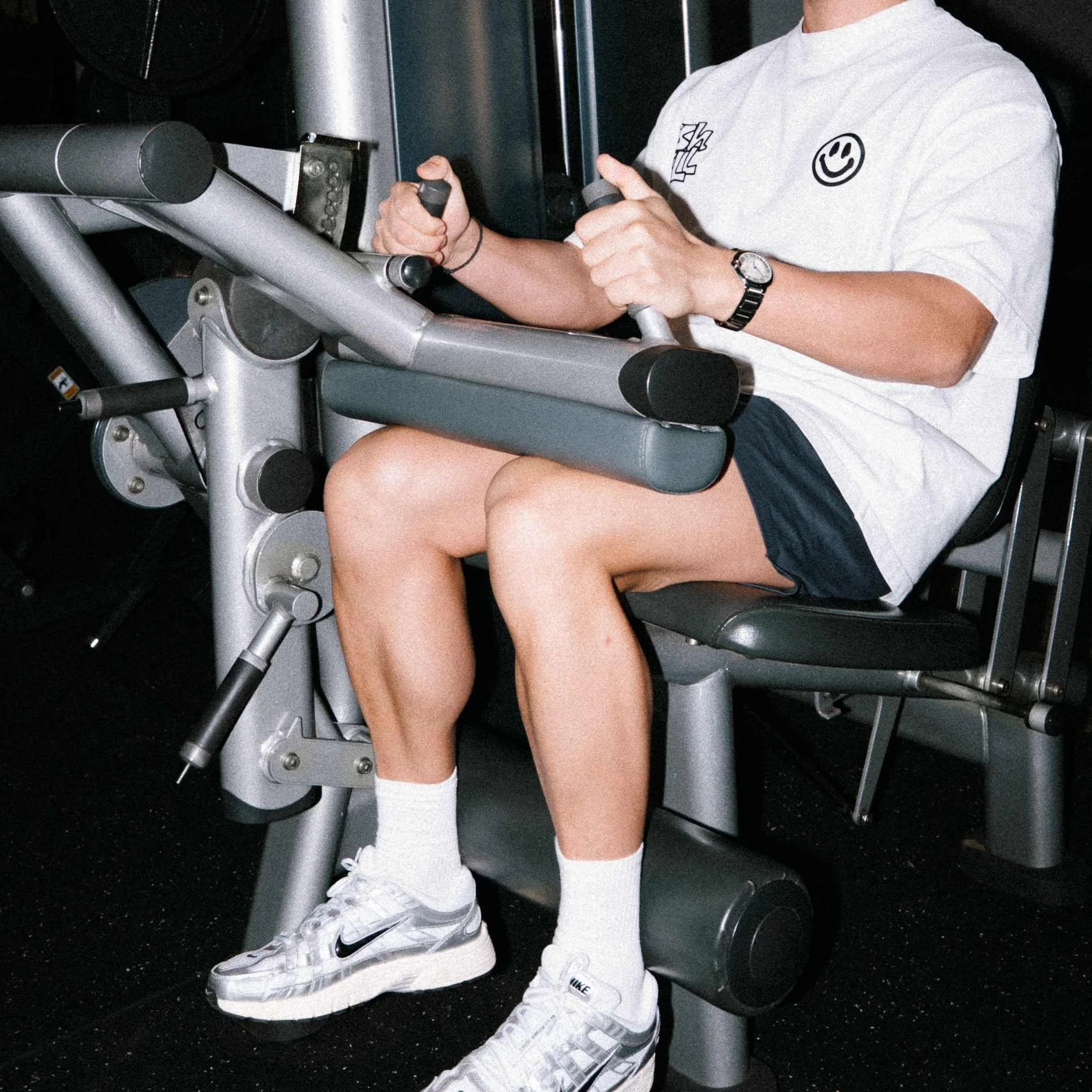 Person riding an exercise bike wearing a gray sweatshirt with a smiley face and skateboard graphic, navy shorts, white socks, and Nike sneakers, in a gym with black flooring.