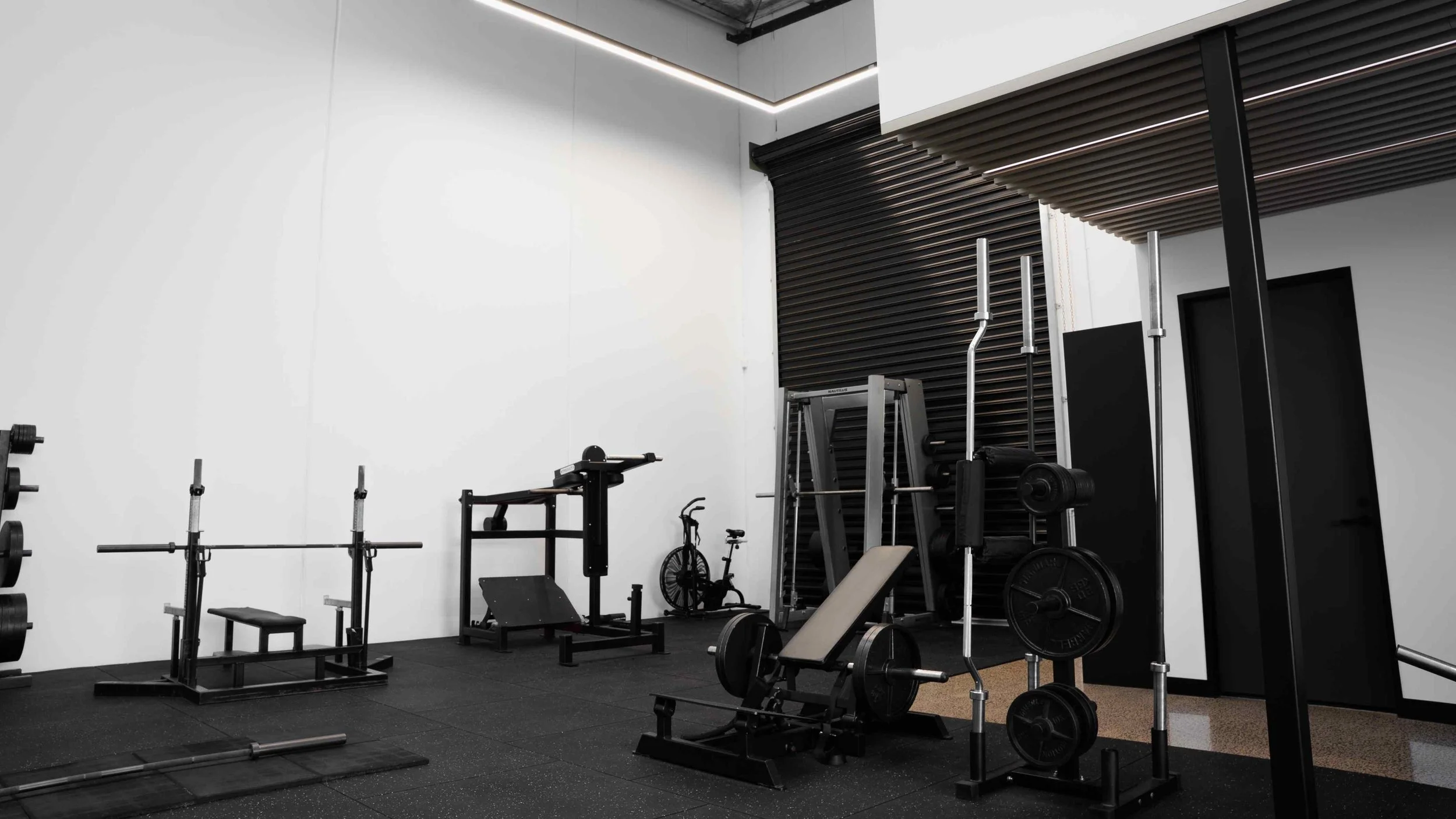 Gym workout area with weightlifting equipment, a stationary bike, and a black roller door in the background.