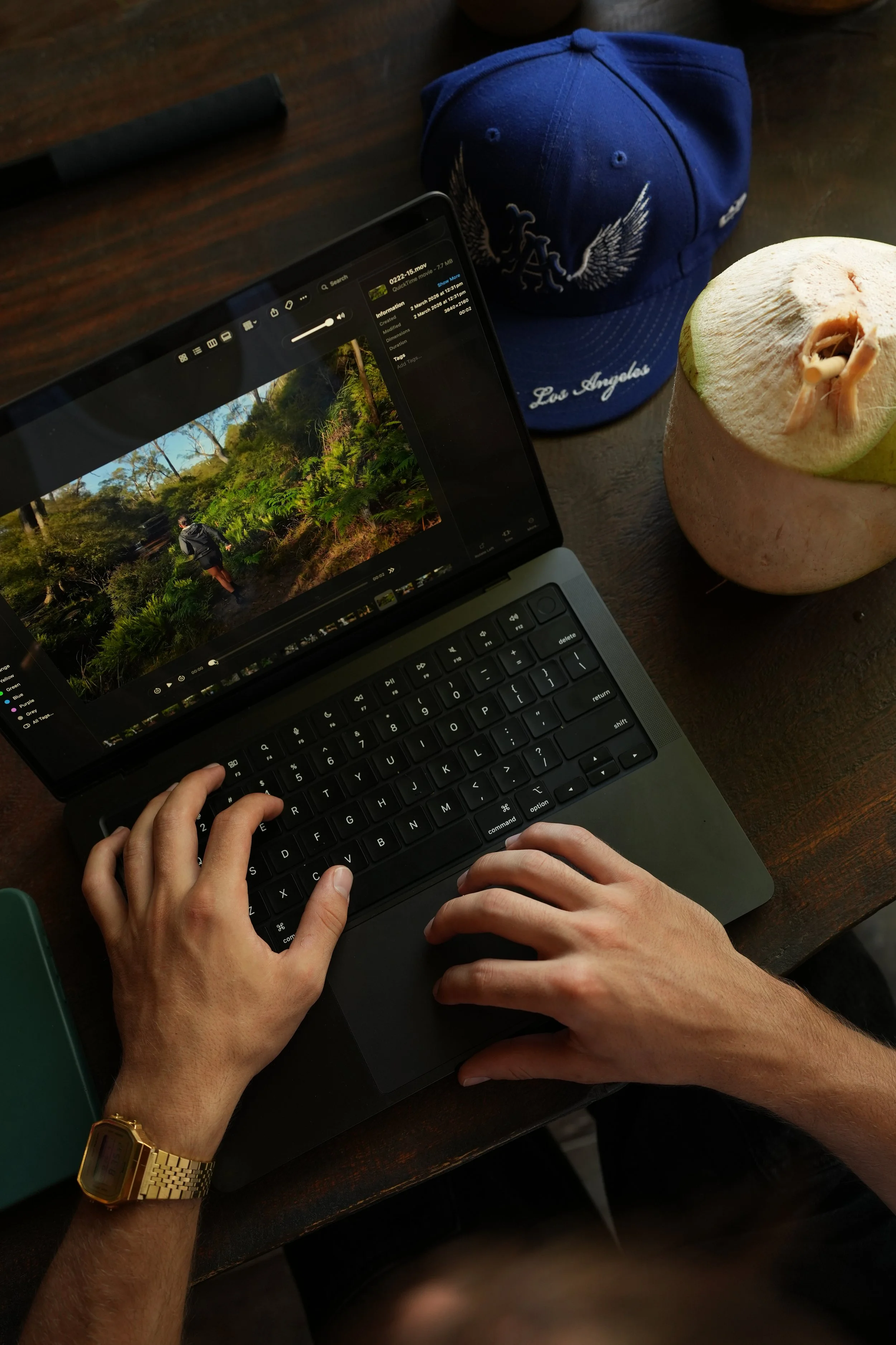 Person typing on a laptop keyboard with hands visible, wearing a gold watch on the left wrist. On the table are a blue Los Angeles cap, a green notebook, a coconut with a straw, and a tablet showing a nature photo.